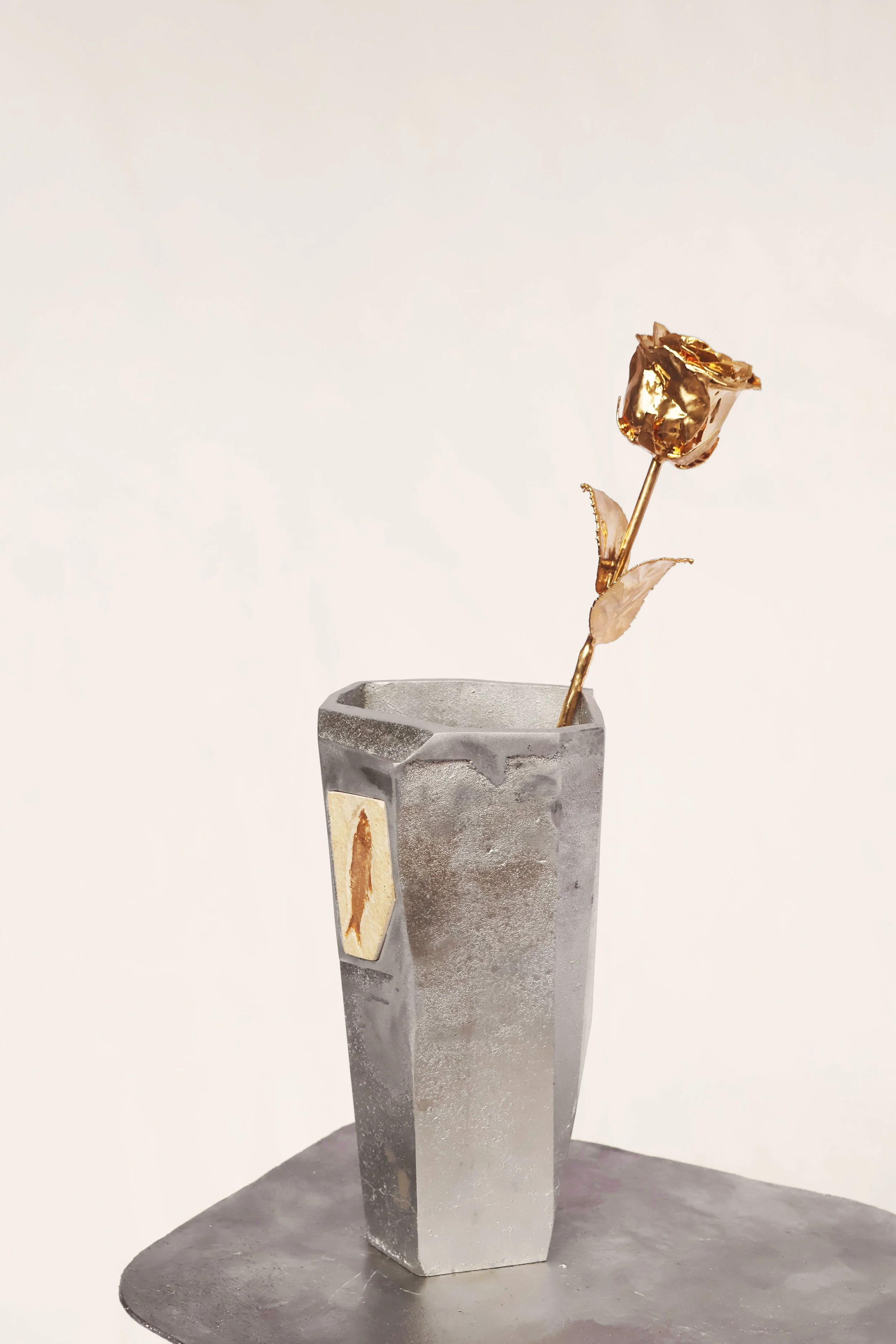A metallic vase with a gold fish emblem contains a single golden rose with a bent stem, set on a gray surface against a plain beige background.