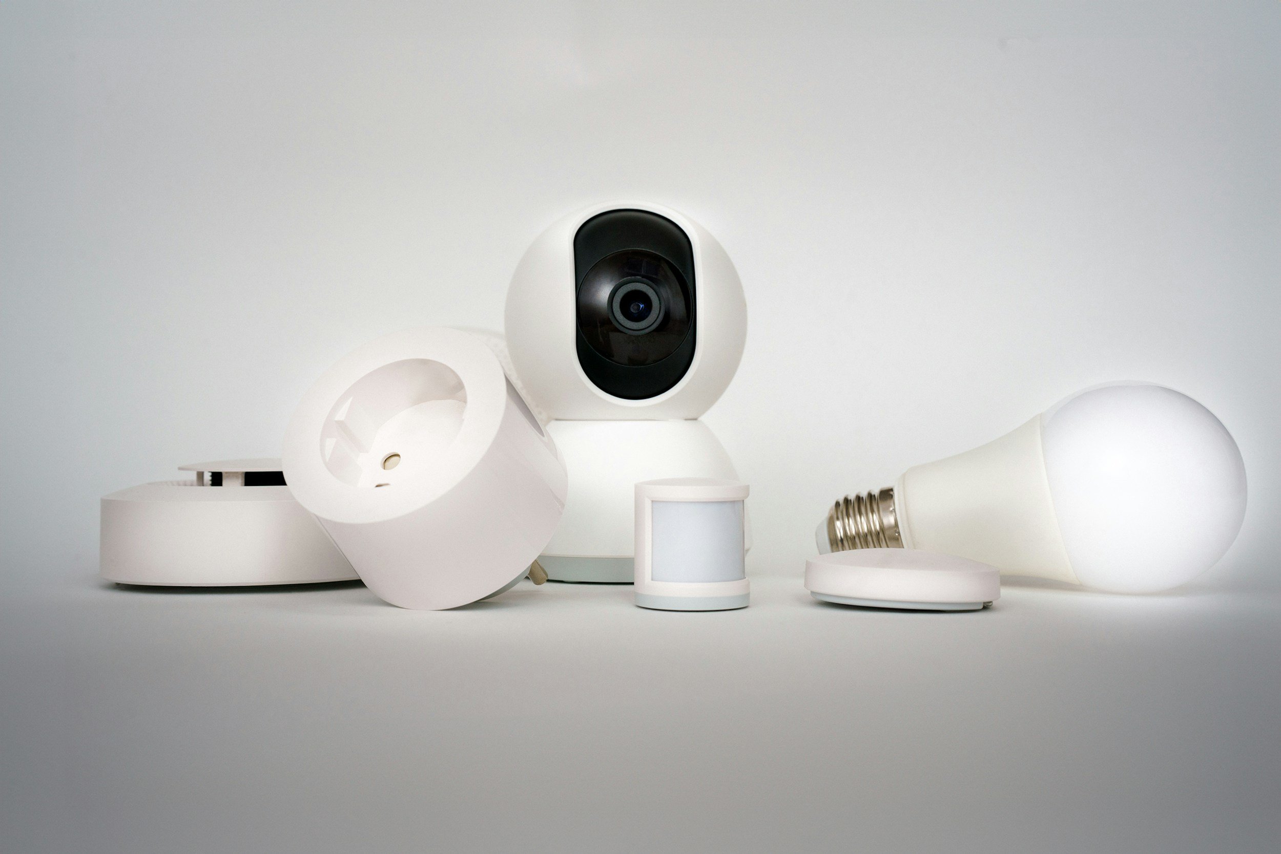 Collection of smart home devices including a security camera, motion sensor, light bulb, and other connected gadgets on a white surface.