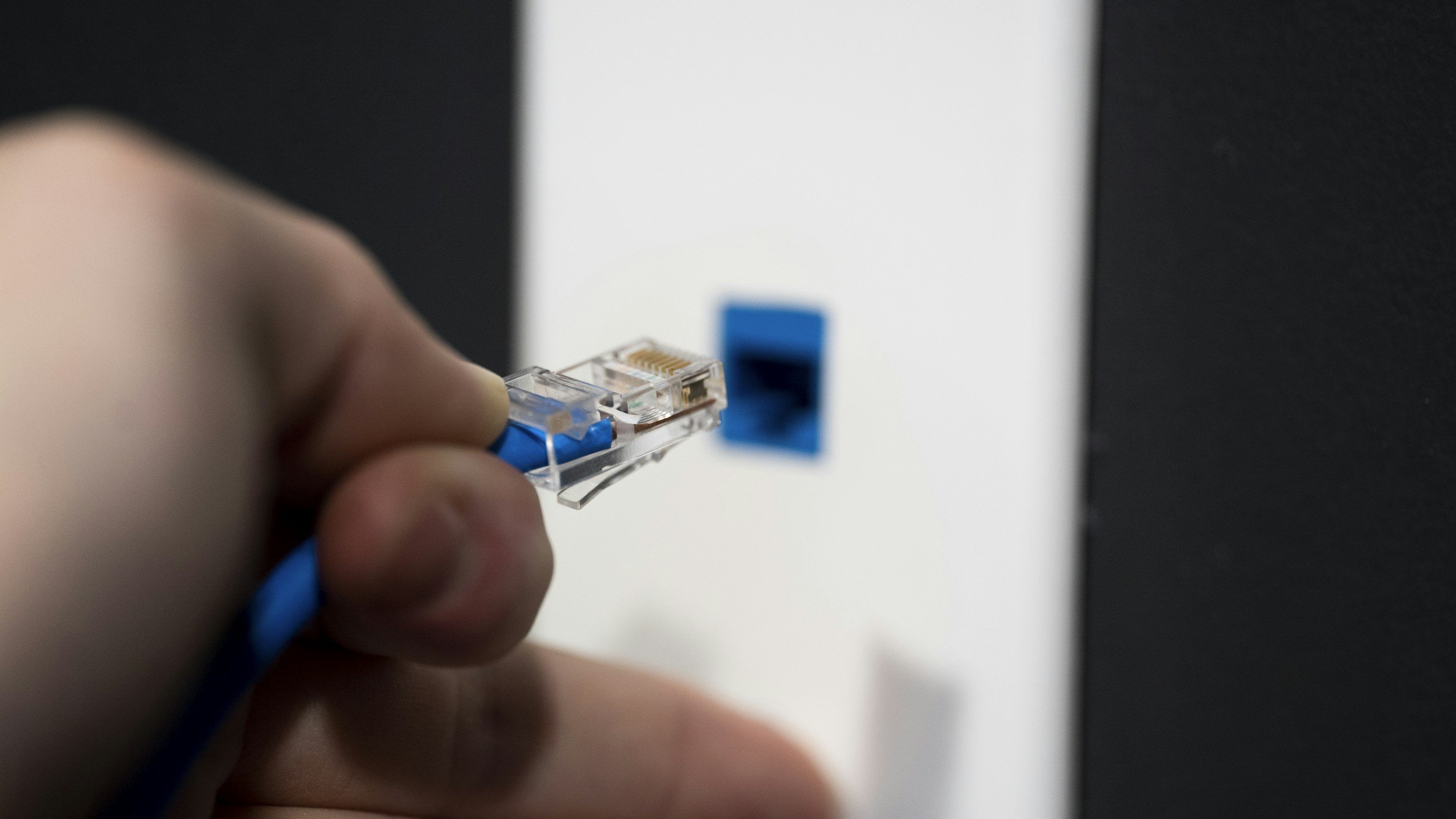 Close-up of a hand holding an Ethernet cable with an RJ45 connector, about to be plugged into a network port on a wall.