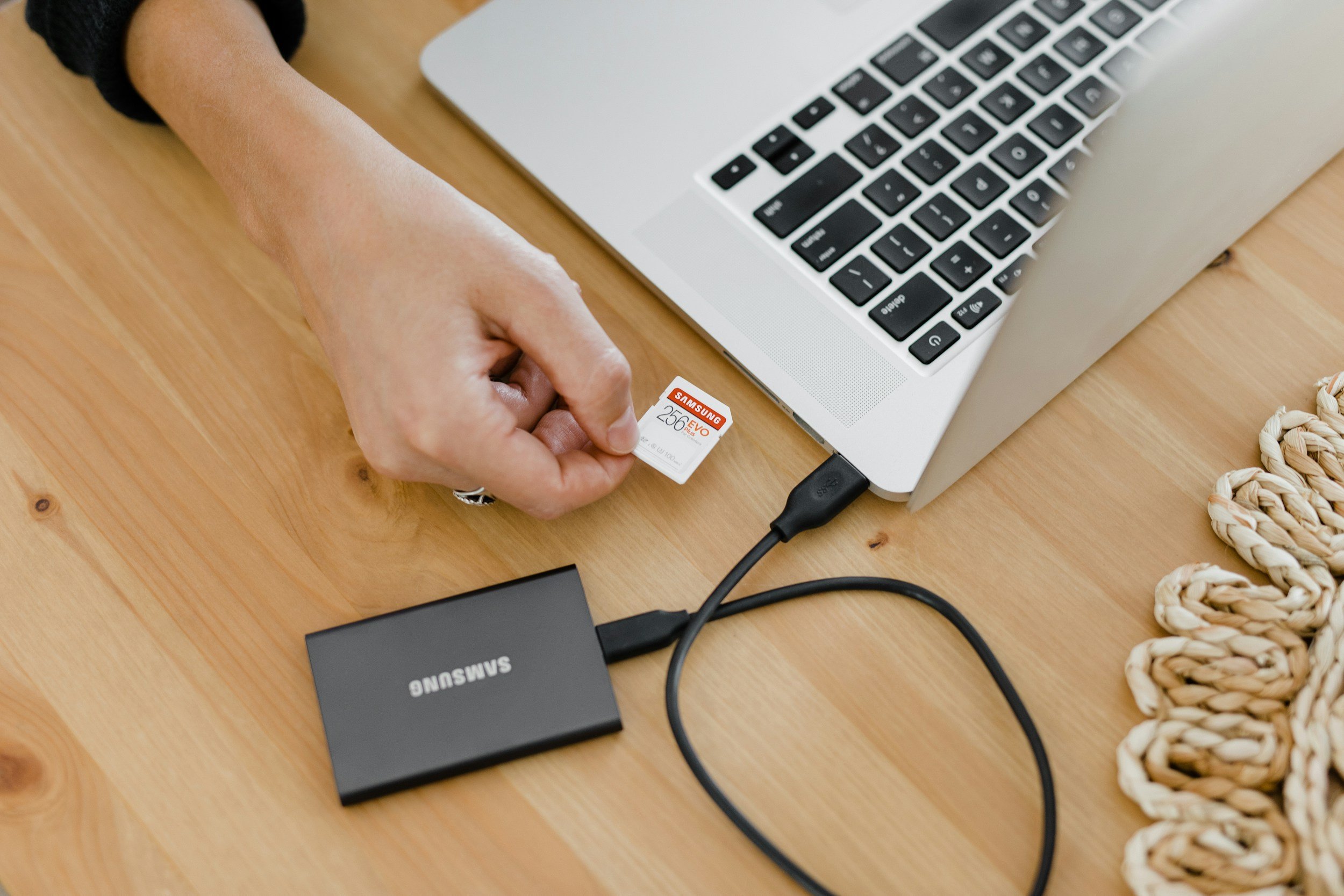 A person inserts a Samsung 256GB EVO SD card into a laptop with an orange wood desk surface. A black Samsung external hard drive and a coiled beige and white woven mat are also on the desk.