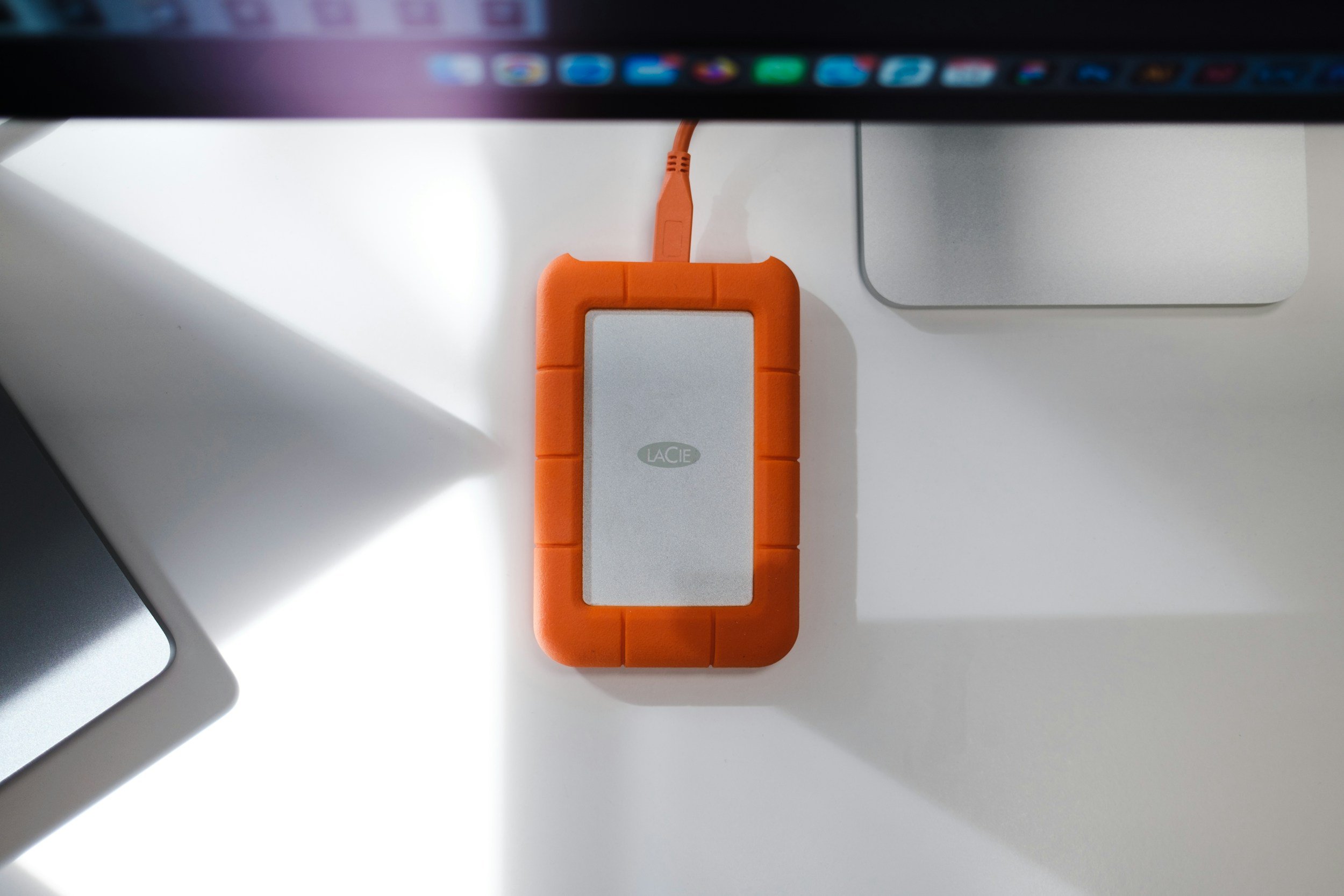 Orange LaCie external hard drive on a white desk, connected by an orange cable, surrounded by a computer monitor and a silver electronic device.