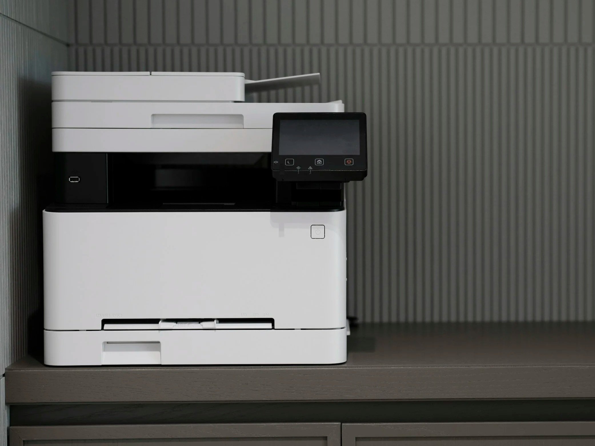 A modern office printer or copier on a wooden table against a gray textured wall.