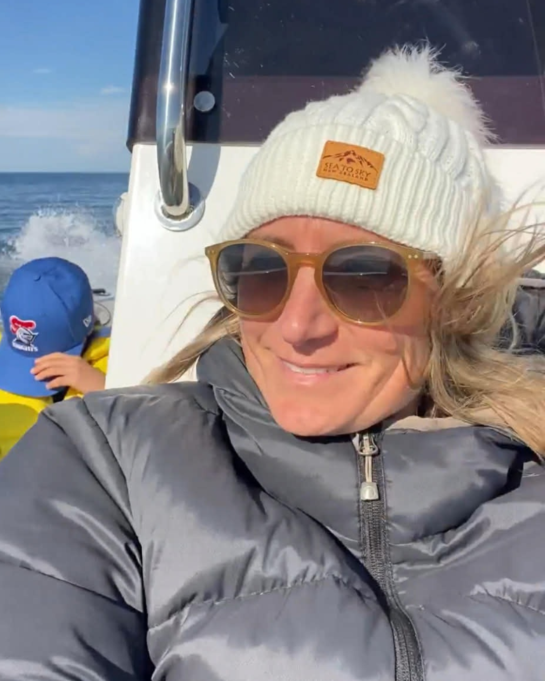 A woman taking a selfie on a boat, wearing a white knit beanie, sunglasses, and a gray jacket; a child in a blue cap and yellow jacket is in the background.