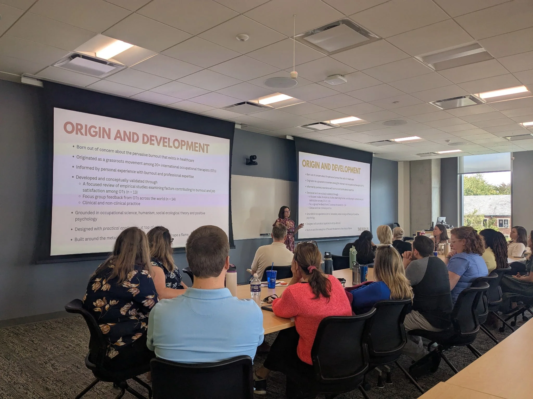 A diverse group of people attending a presentation in a conference room. A woman speaker stands in front of a large screen displaying a slide titled 'Origin and Development'. The room is well-lit with large windows on the right side, showing a view of trees and a building outside.