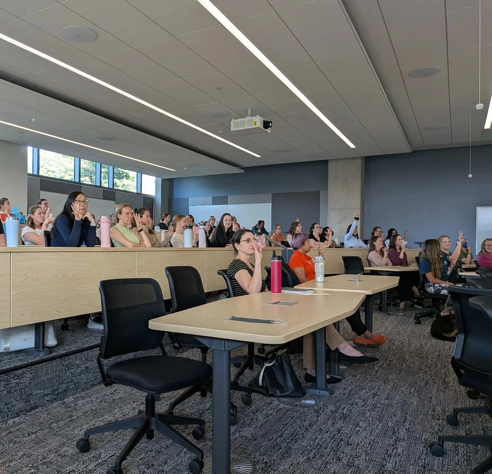 A large classroom or conference room filled with students or attendees seated at desks, some raising their hands. The room has high ceilings with linear lighting, large windows on one side, and a modern design.