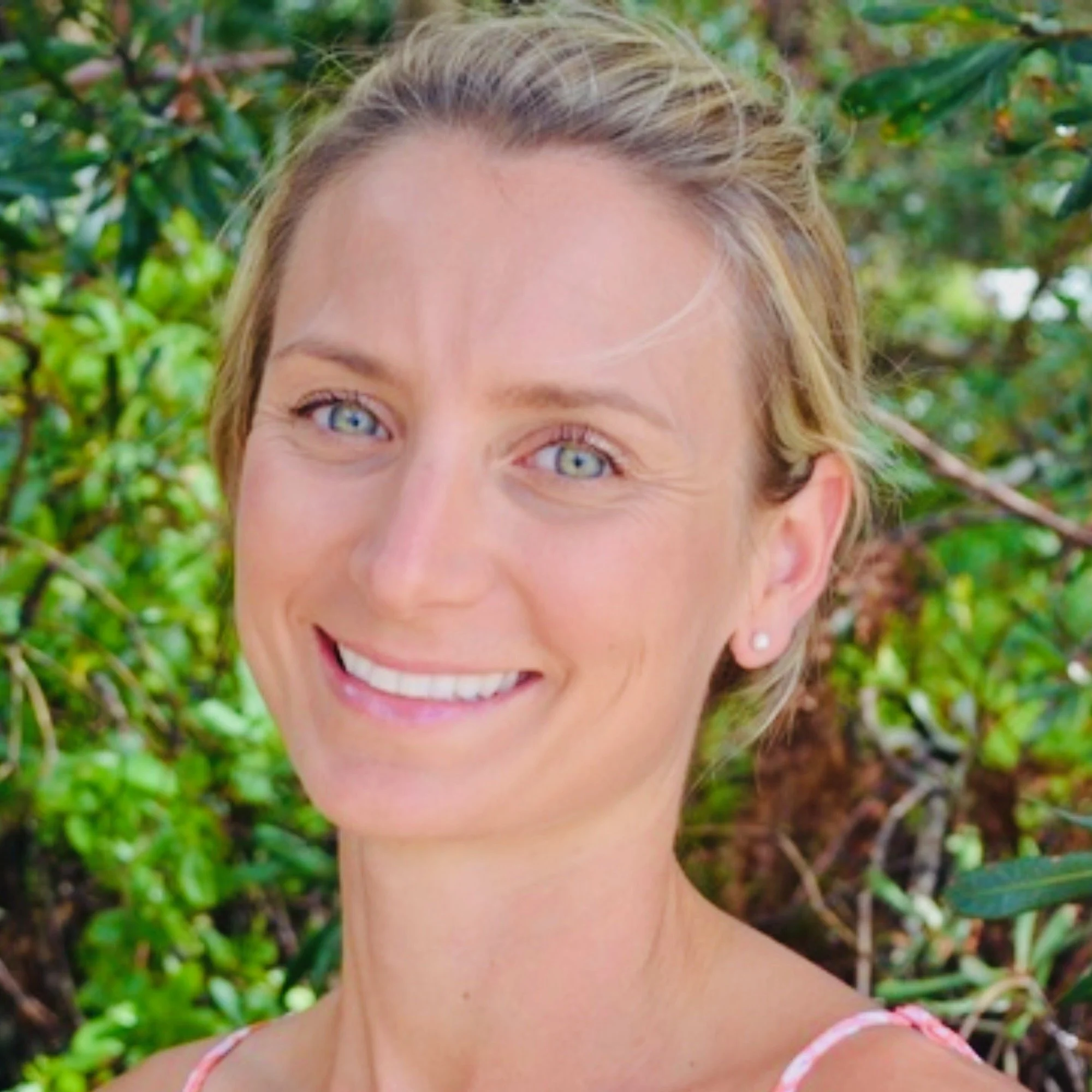 Close-up of a woman with blonde hair, blue eyes, and a bright smile, outdoors with green foliage in the background.