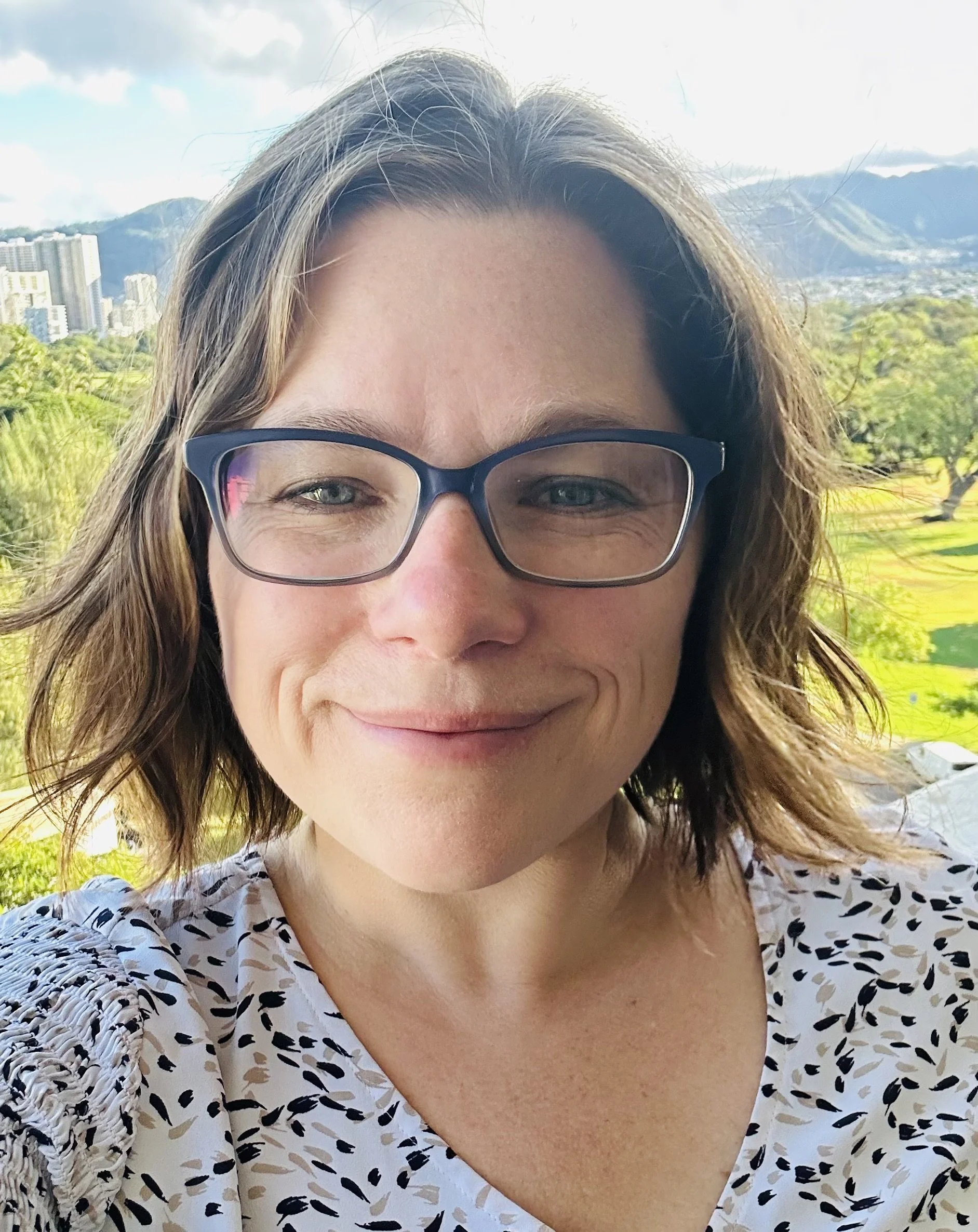 A woman smiling outdoors with glasses, green landscape, trees, city buildings, mountains, and blue sky in the background.