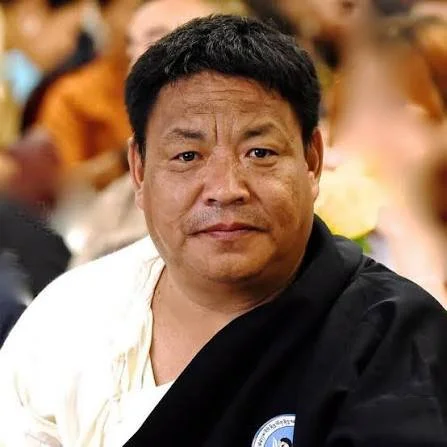 A middle-aged man with short black hair, wearing a black and white outfit, sitting in a crowded indoor setting.
