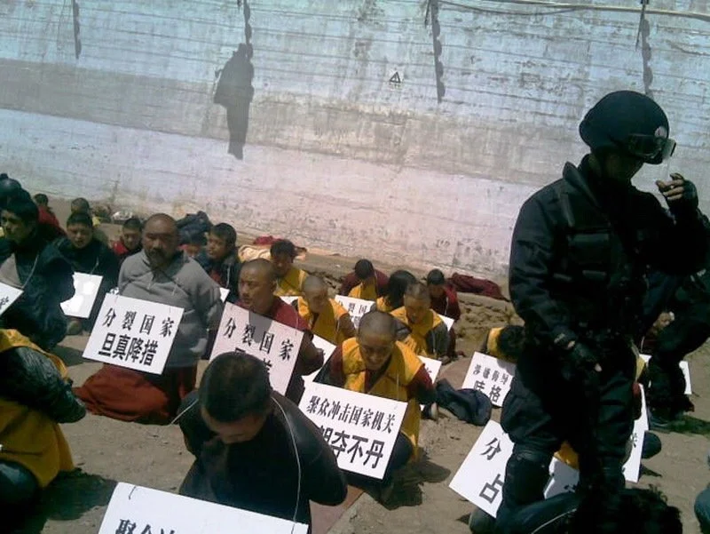 A group of people sitting on the ground against a wall, holding signs with Chinese characters, with a police officer standing nearby wearing protective gear and sunglasses.