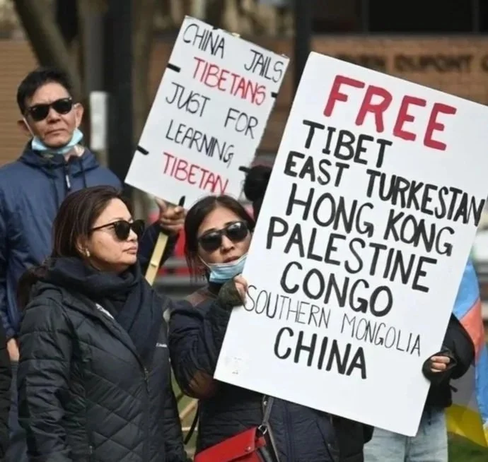 Protesters holding signs criticizing China's treatment of Tibet and other regions, with one sign reading 'FREE Tibet East Turkestan Hong Kong Palestine Congo Southern Mongolia China' and another mentioning 'China Jails Tibetans just for learning Tibet'.