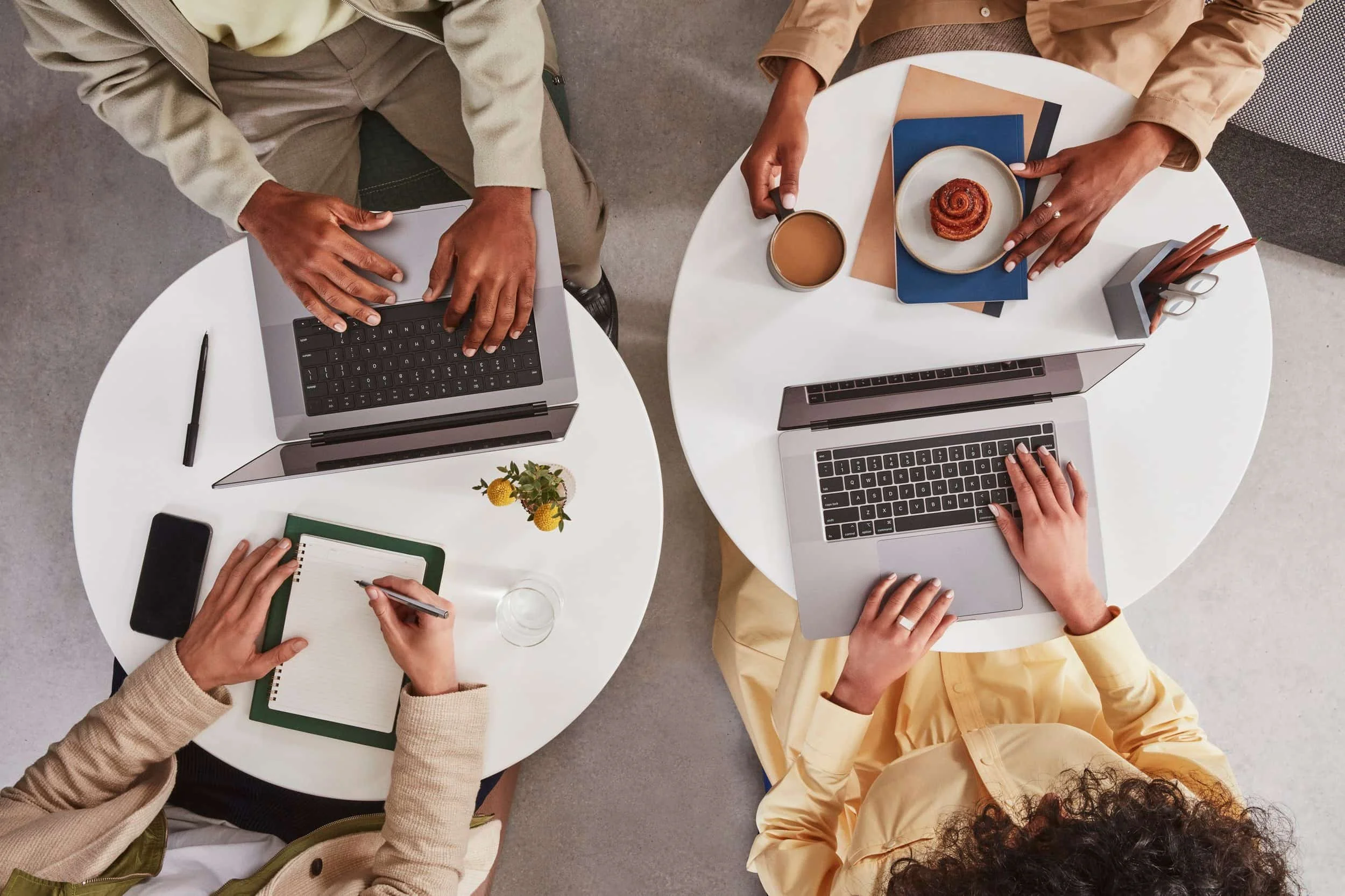 Trois personnes travaillant à des tables rondes avec des ordinateurs portables, un carnet, un stylo, une tasse de café, et un croissant, dans un environnement de bureau.
