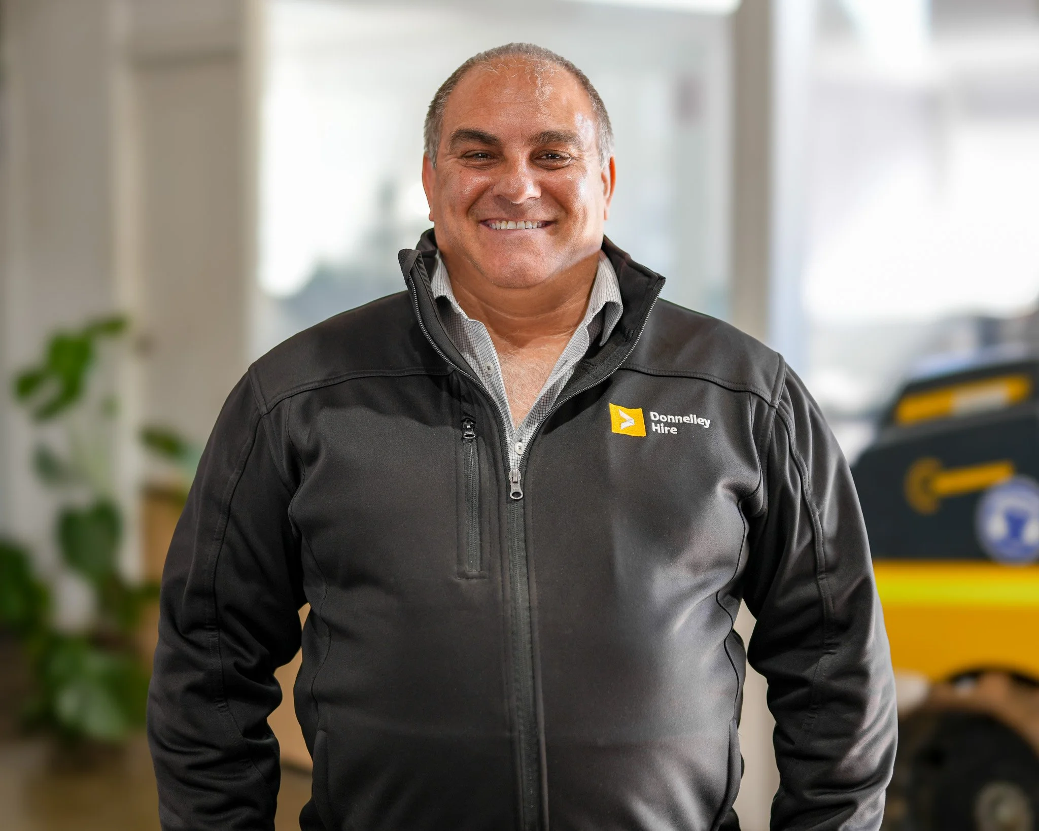 A smiling man wearing a black jacket with a 'Donnelley Hire' logo stands indoors with blurred background of offices and equipment.