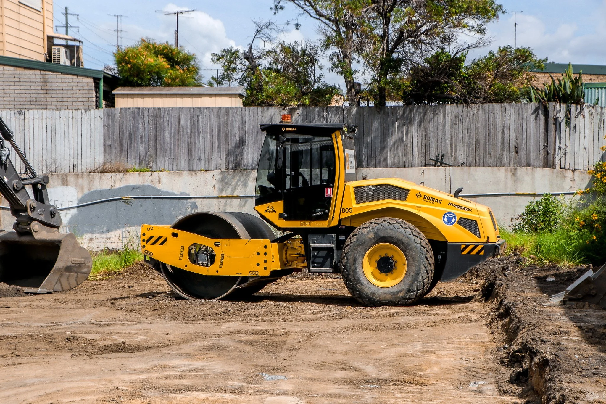 Yellow road roller at a construction site with dirt, a wooden fence, trees, and buildings in the background.