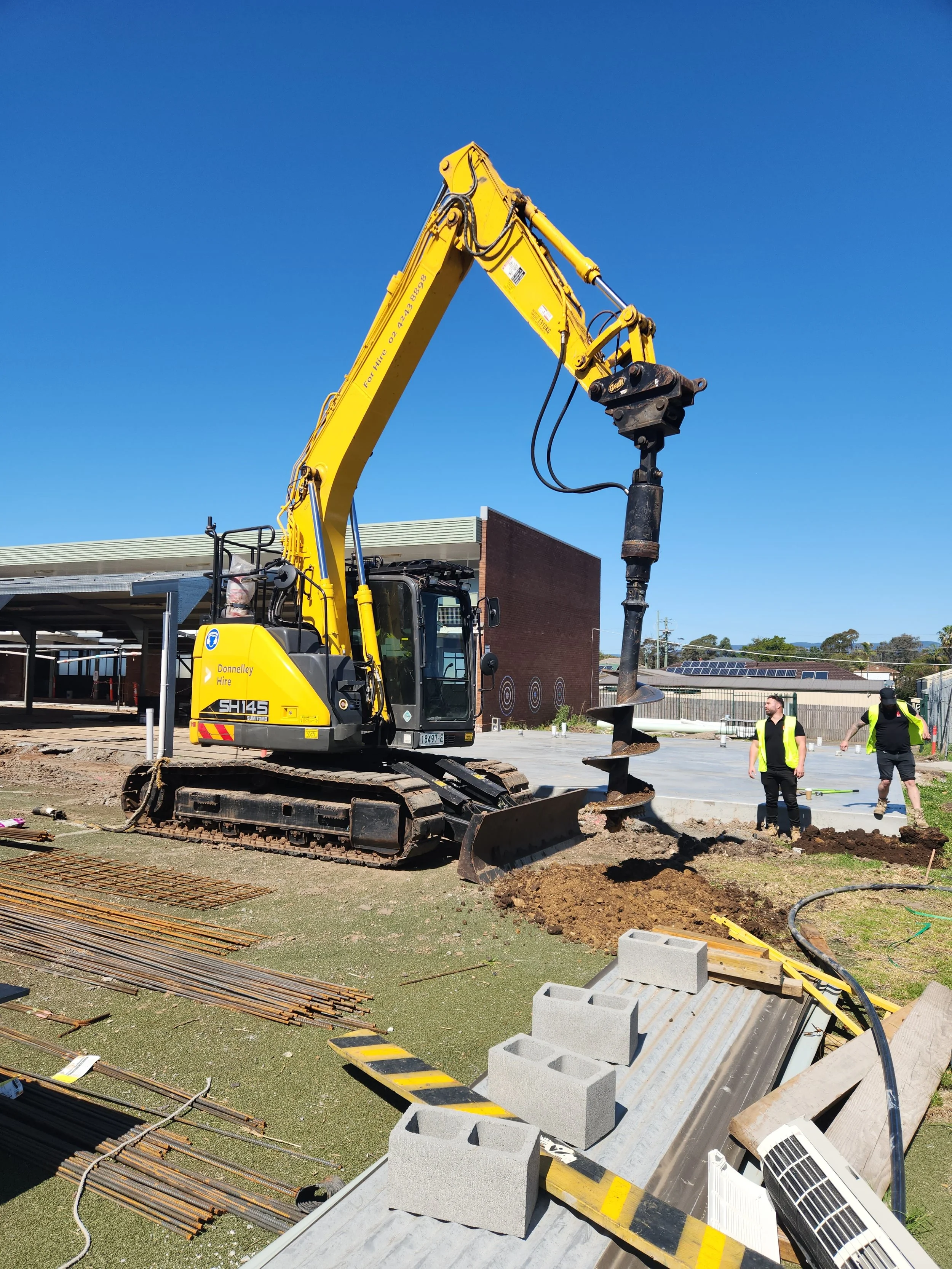 Close-up of a hydraulic attachment on a construction excavator, with yellow machinery and green trees in the background.