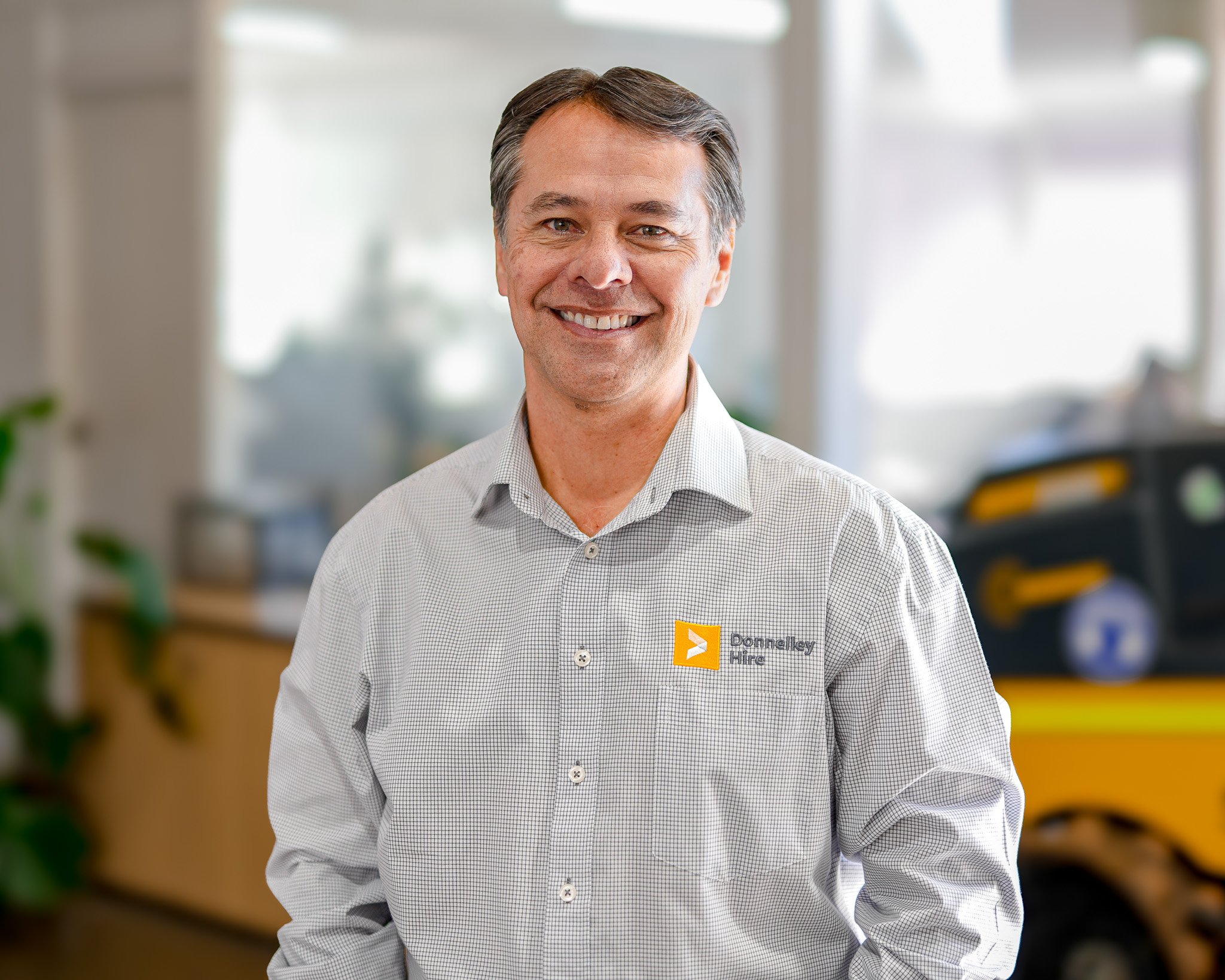 A smiling man wearing a light gray checkered shirt with a Dannelkey Hire logo stands indoors with a blurred background, including a yellow and black machine and some office furniture.