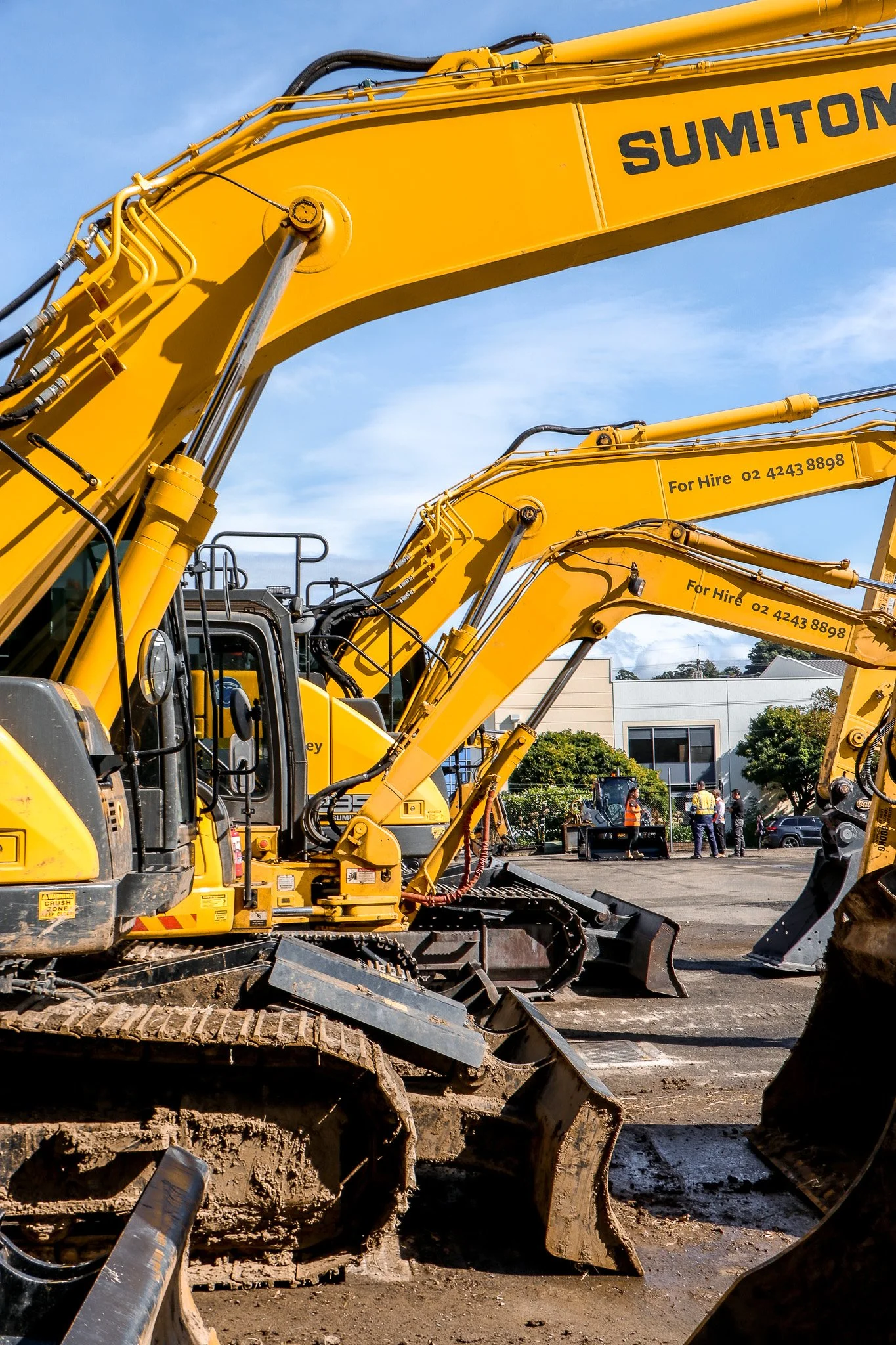Multiple yellow excavators parked on a construction site with blue sky and buildings in the background.