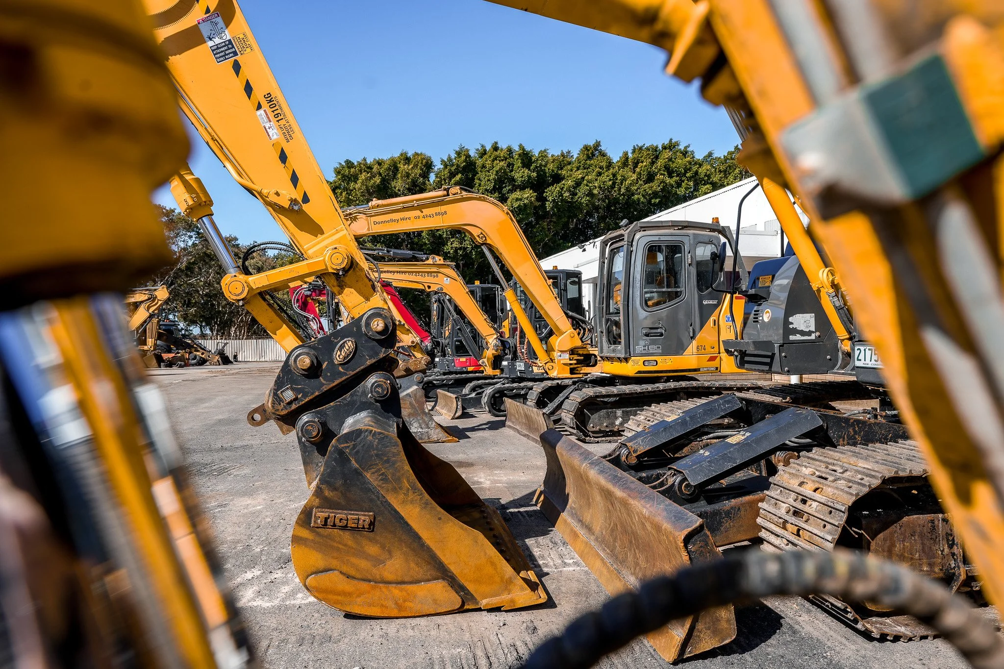 Multiple yellow and black construction excavators parked on a dirt lot with green trees and white industrial buildings in the background.