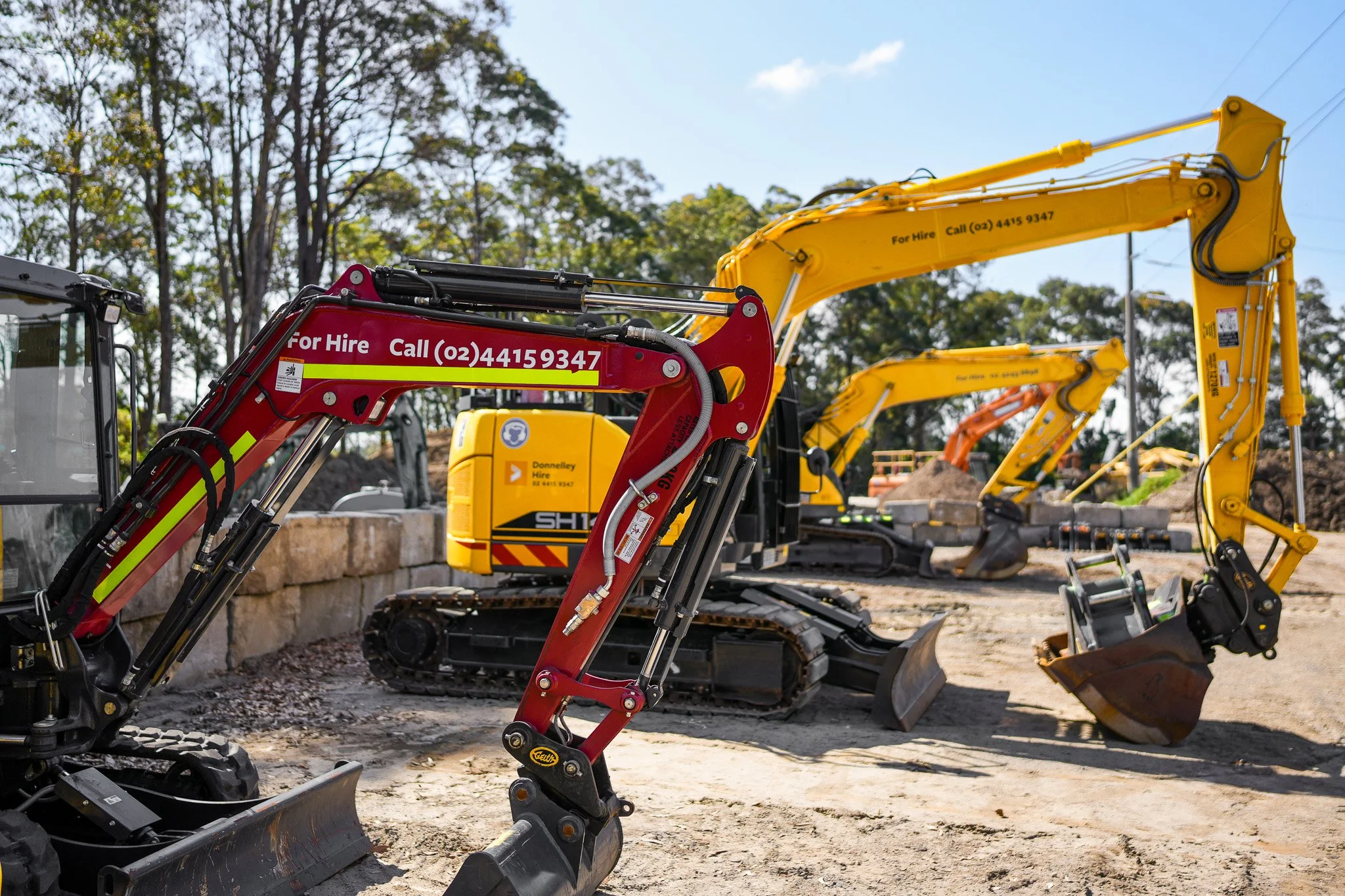 Construction site with multiple excavators and machinery, some with 'For Hire' and contact number banners.