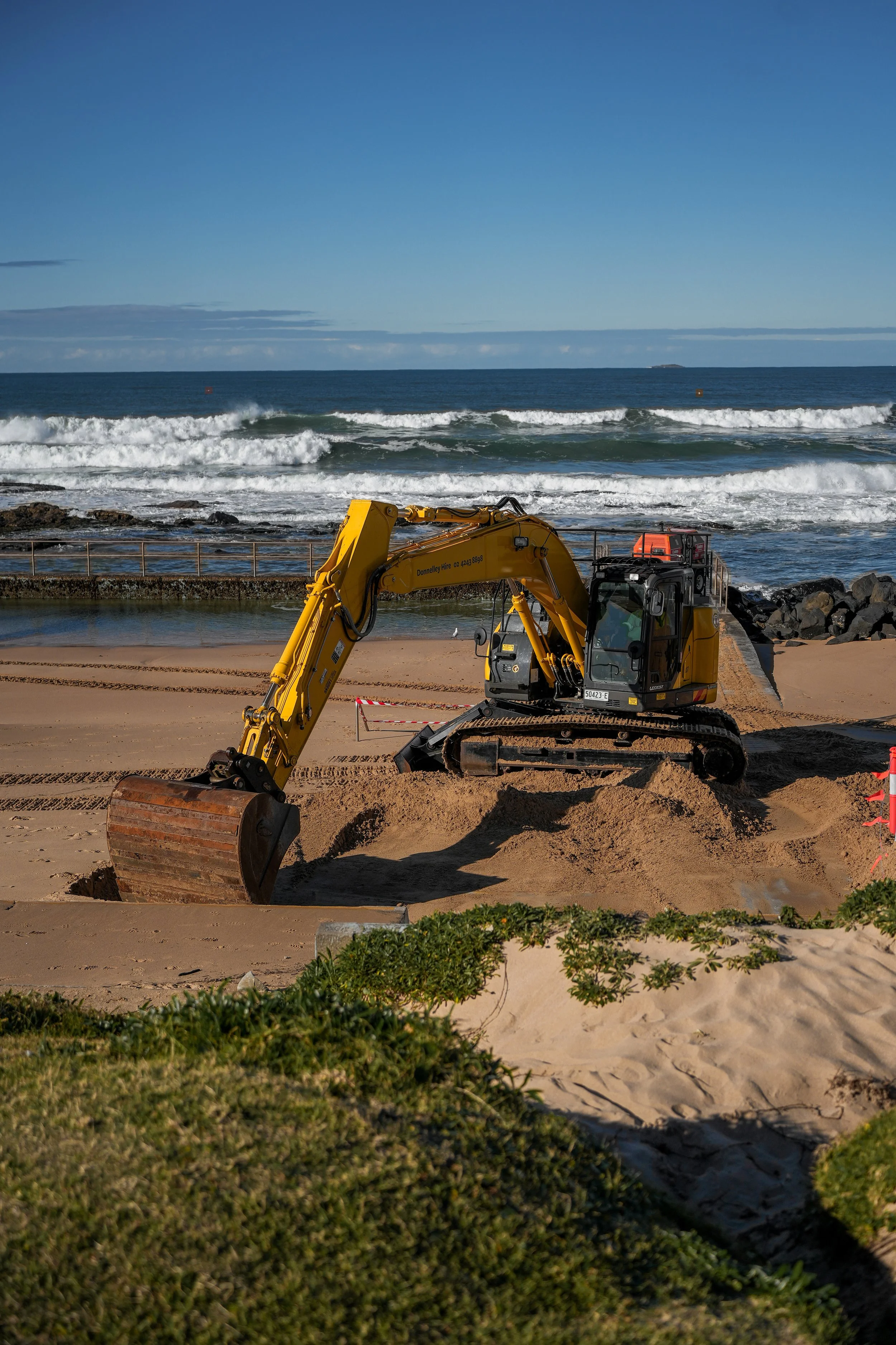 Yellow excavator digging a hole on a sandy beach with the ocean and city skyline in the background.