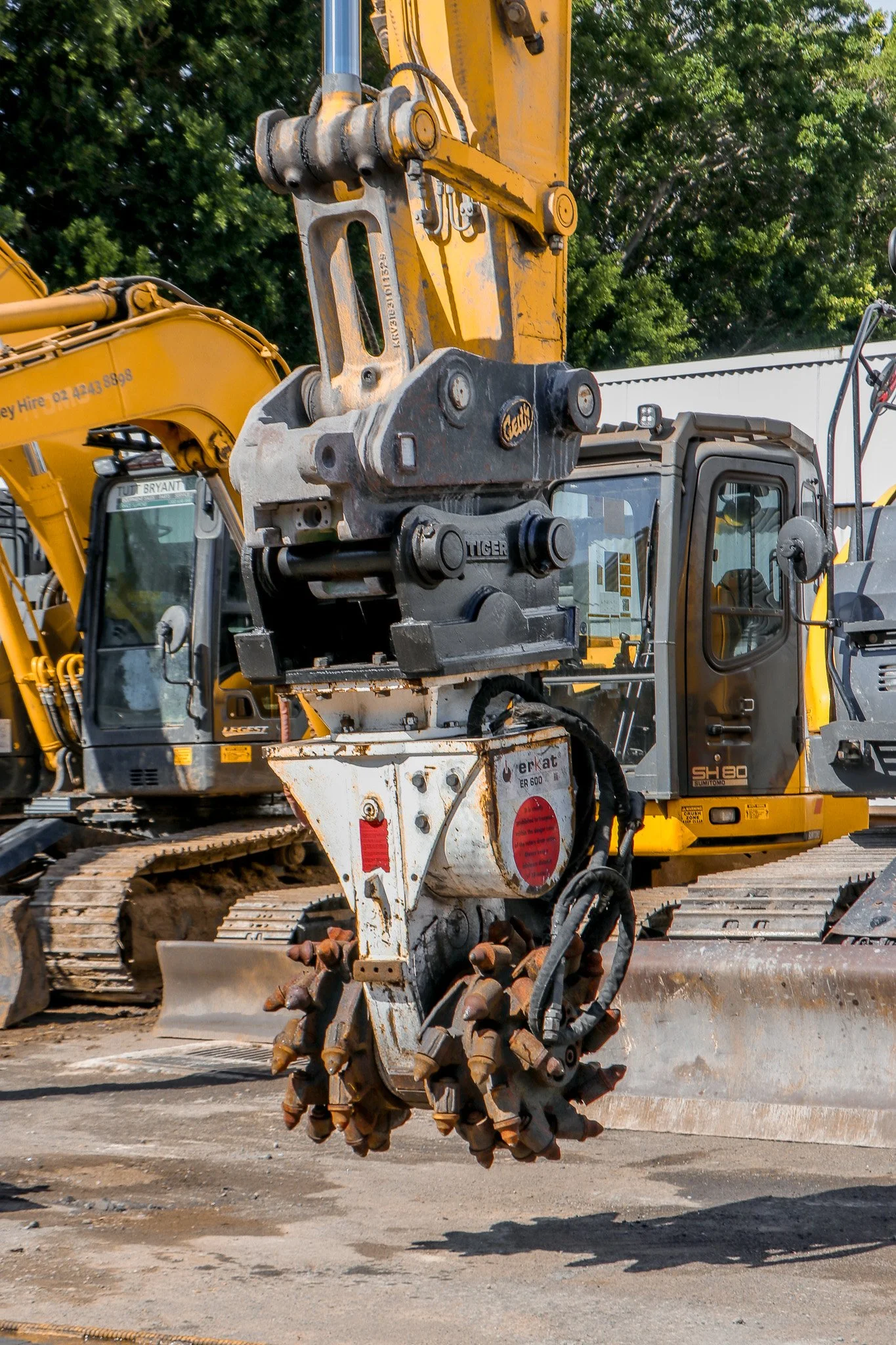 Close-up of a hydraulic demolition attachment with rotating sprockets, attached to an excavator at a construction site, with other construction equipment in the background.