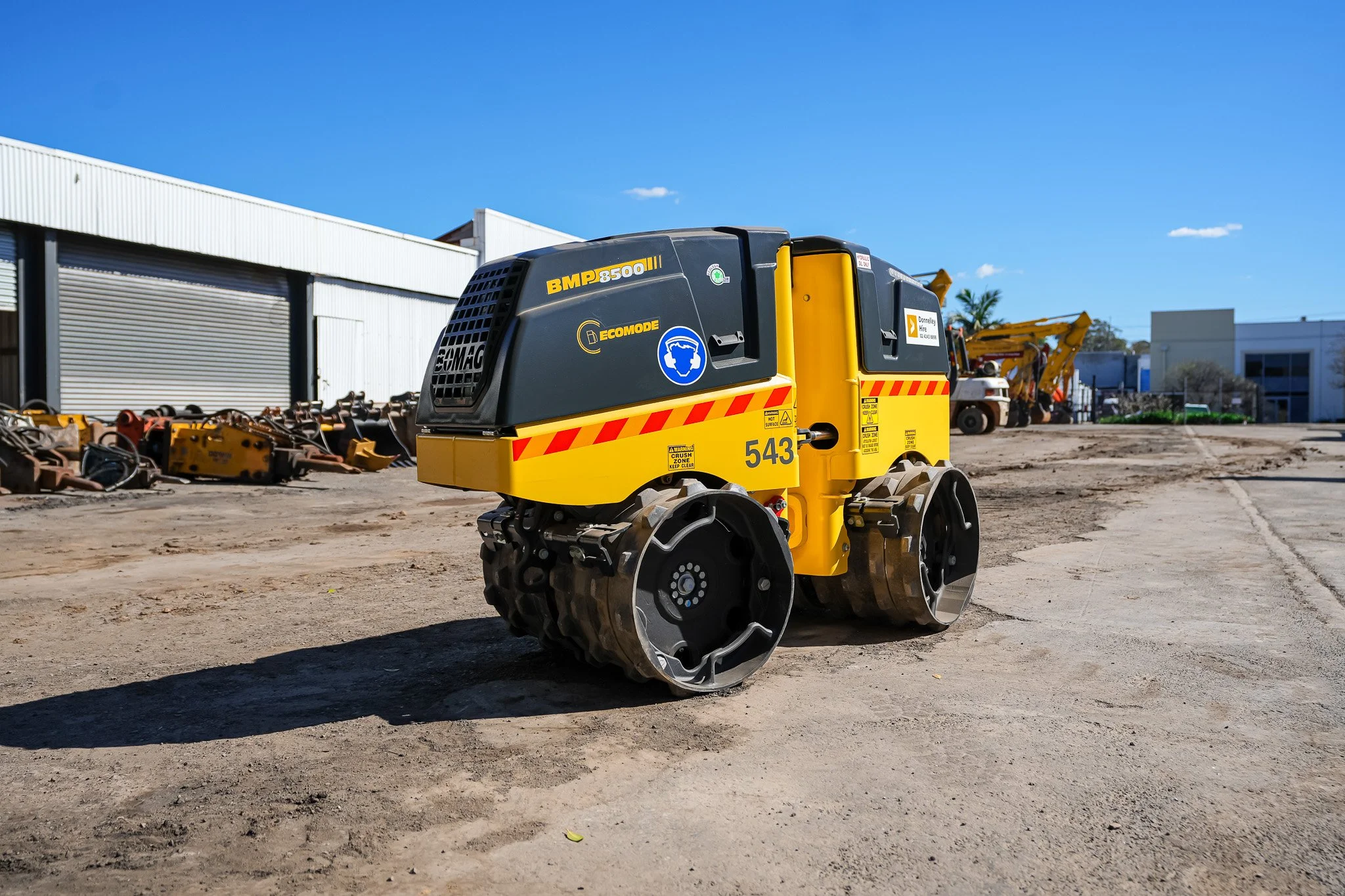 A small yellow and black vibratory compactor machine is parked outdoors on a dirt surface near a workshop with metal doors and other construction equipment in the background.