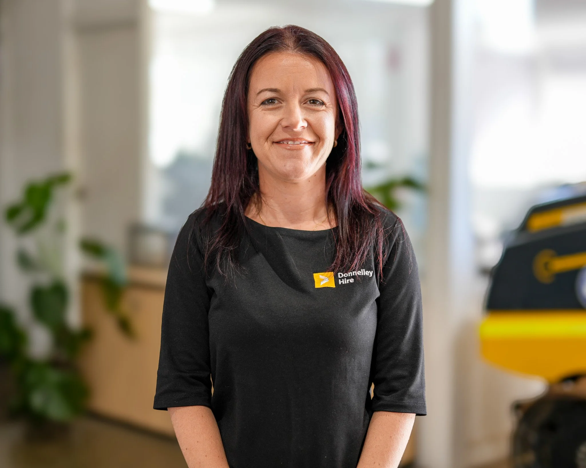 A woman with dark red hair and a black shirt smiling, standing inside an office with plants and yellow equipment in the background.