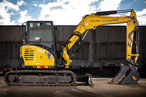 Yellow and black compact excavator on construction site with a concrete wall in the background.