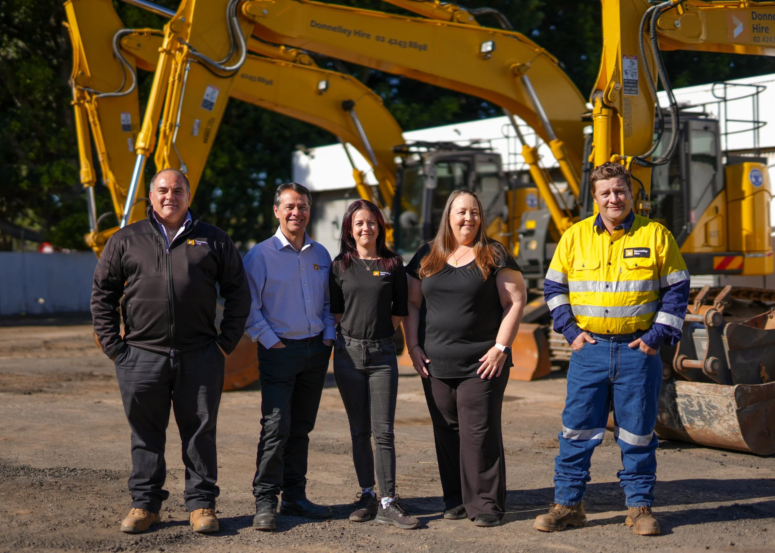Five people standing in front of construction equipment, with two large yellow excavators in the background. They are outdoors on a construction site.