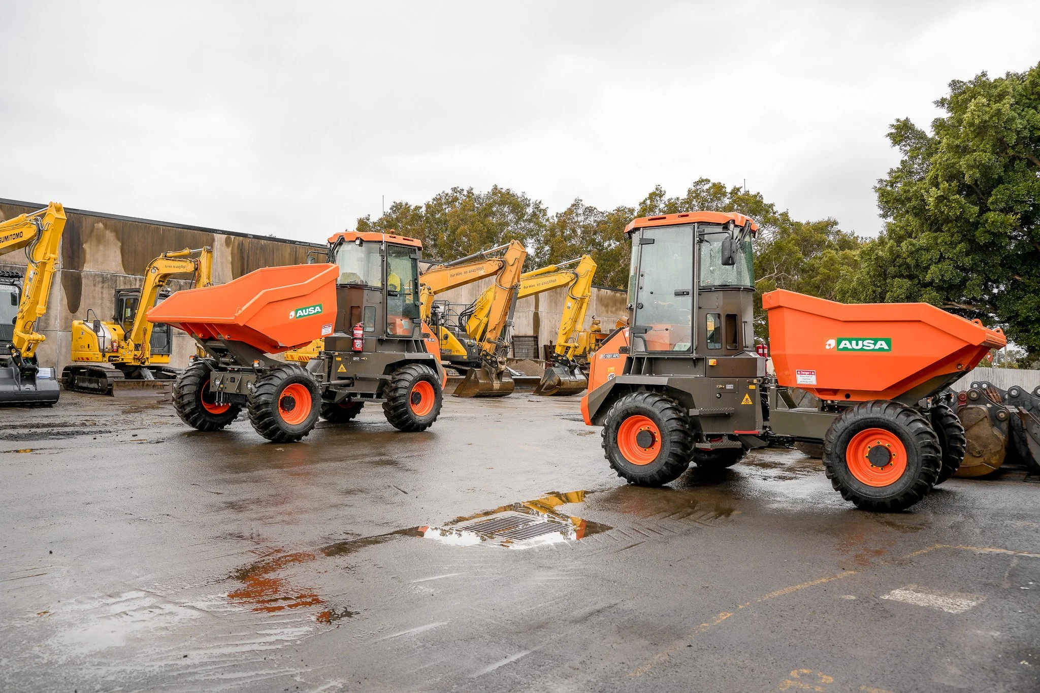 Construction vehicles parked outdoors, including an orange and black dumper and multiple yellow excavators with long arms and buckets, on a wet surface with a concrete wall and trees in the background.