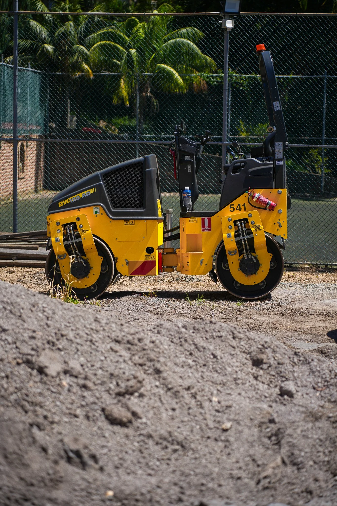 A yellow road roller on a construction site gravel surface with a chain link fence and green trees in the background.