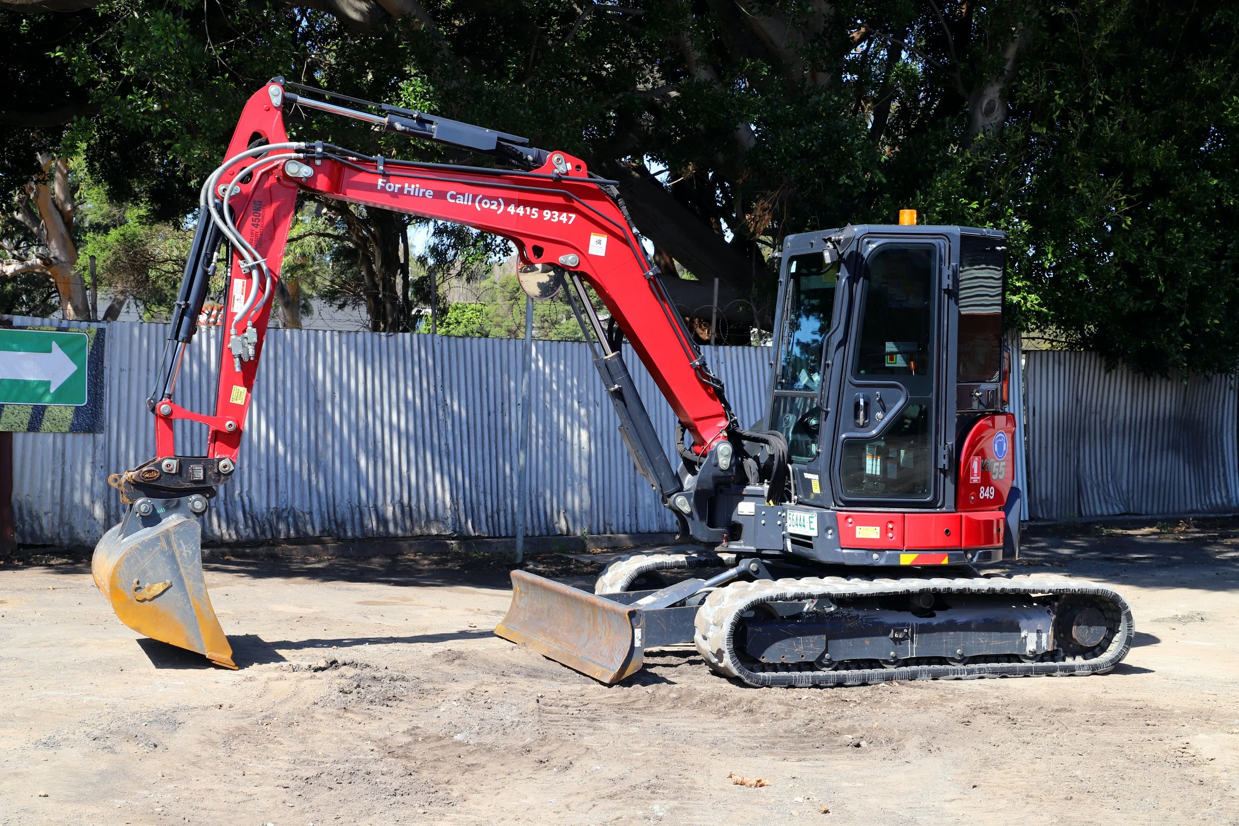 Yellow and black compact excavator on a gravel surface with a dark wooden wall in the background.