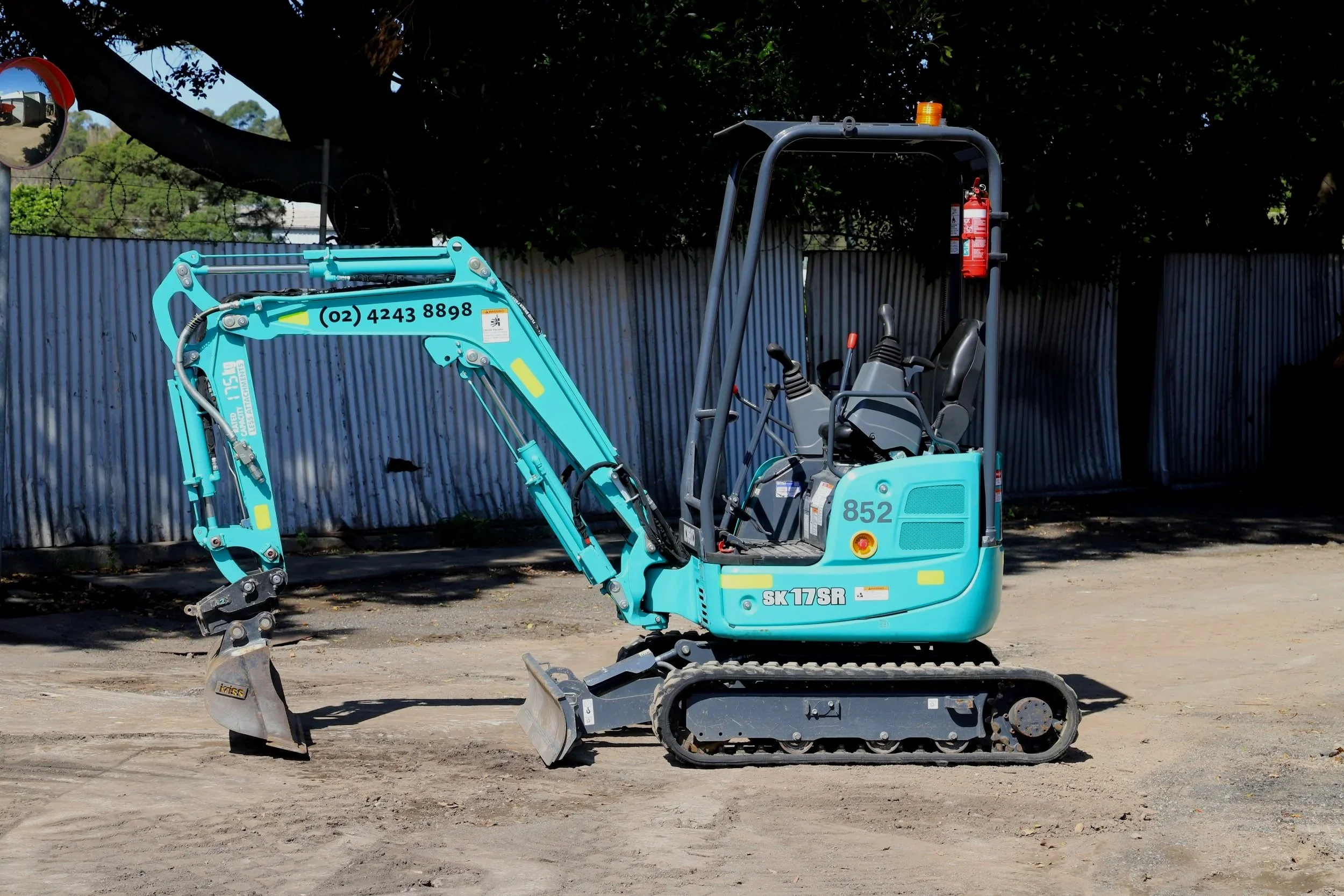 Small teal excavator with black tracks and a black blade, branded 'Kobelco' and 'SK 17SR', positioned against a blue gradient background.