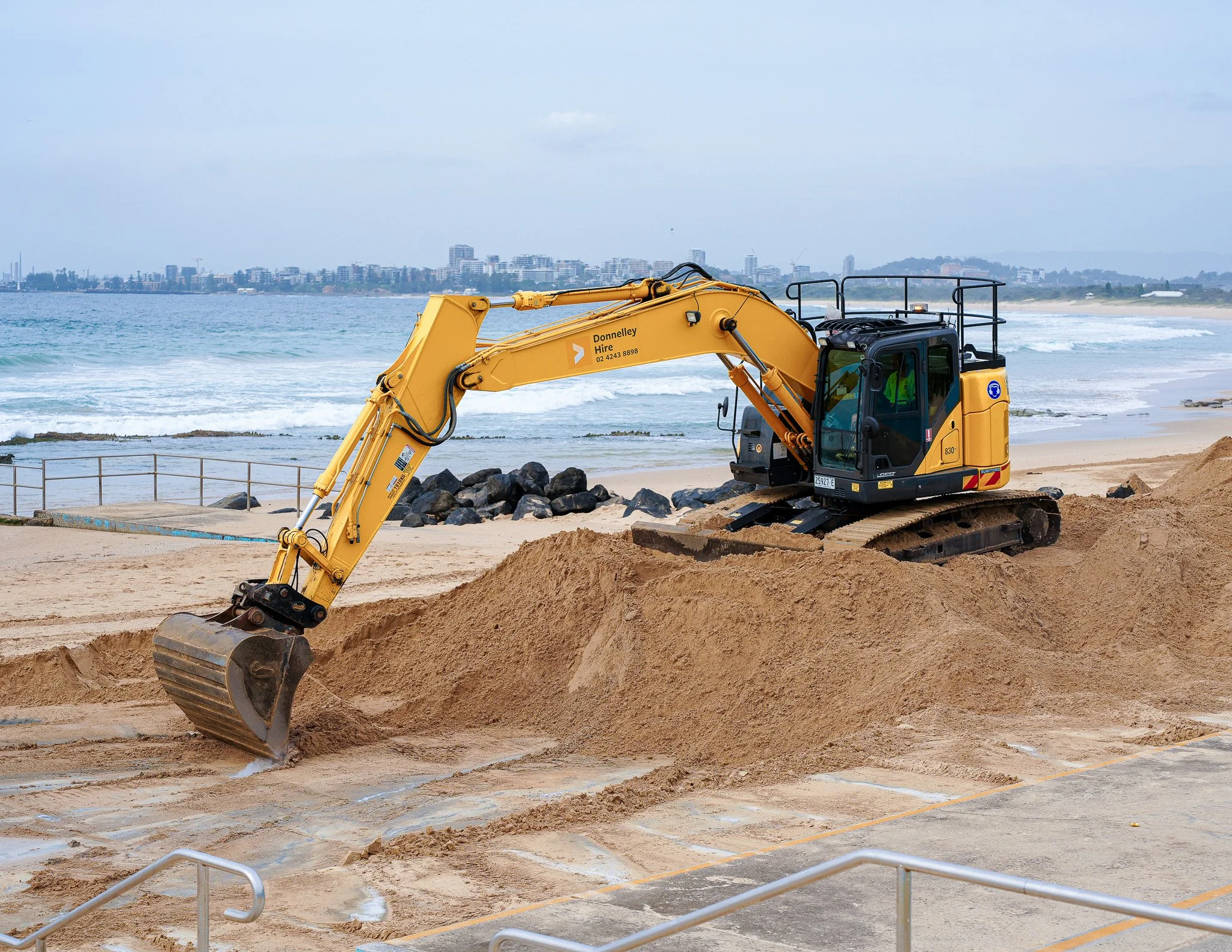 Yellow excavator working on a sandy beach near the ocean with a city skyline in the background.
