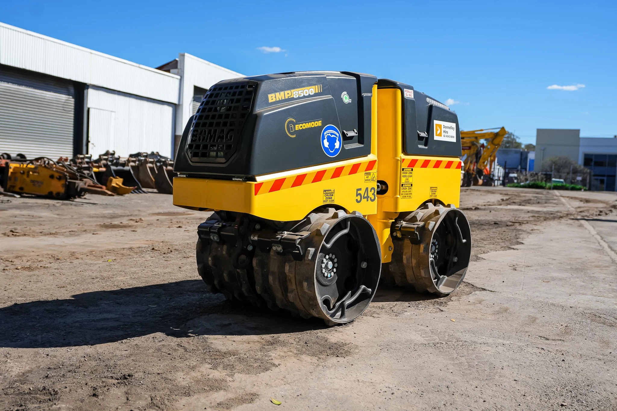 Yellow and black compact construction roller with rubber wheels on dirt ground, with industrial buildings in the background.