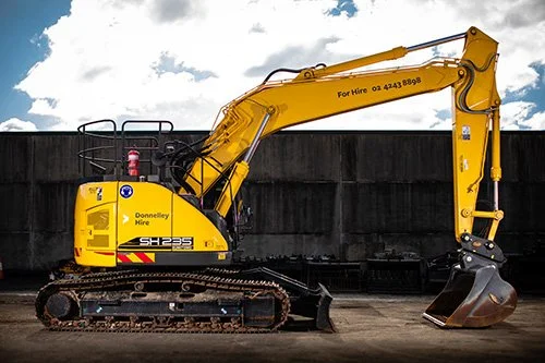 Yellow excavator with black tracks parked outdoors against a dark wall and cloudy sky.
