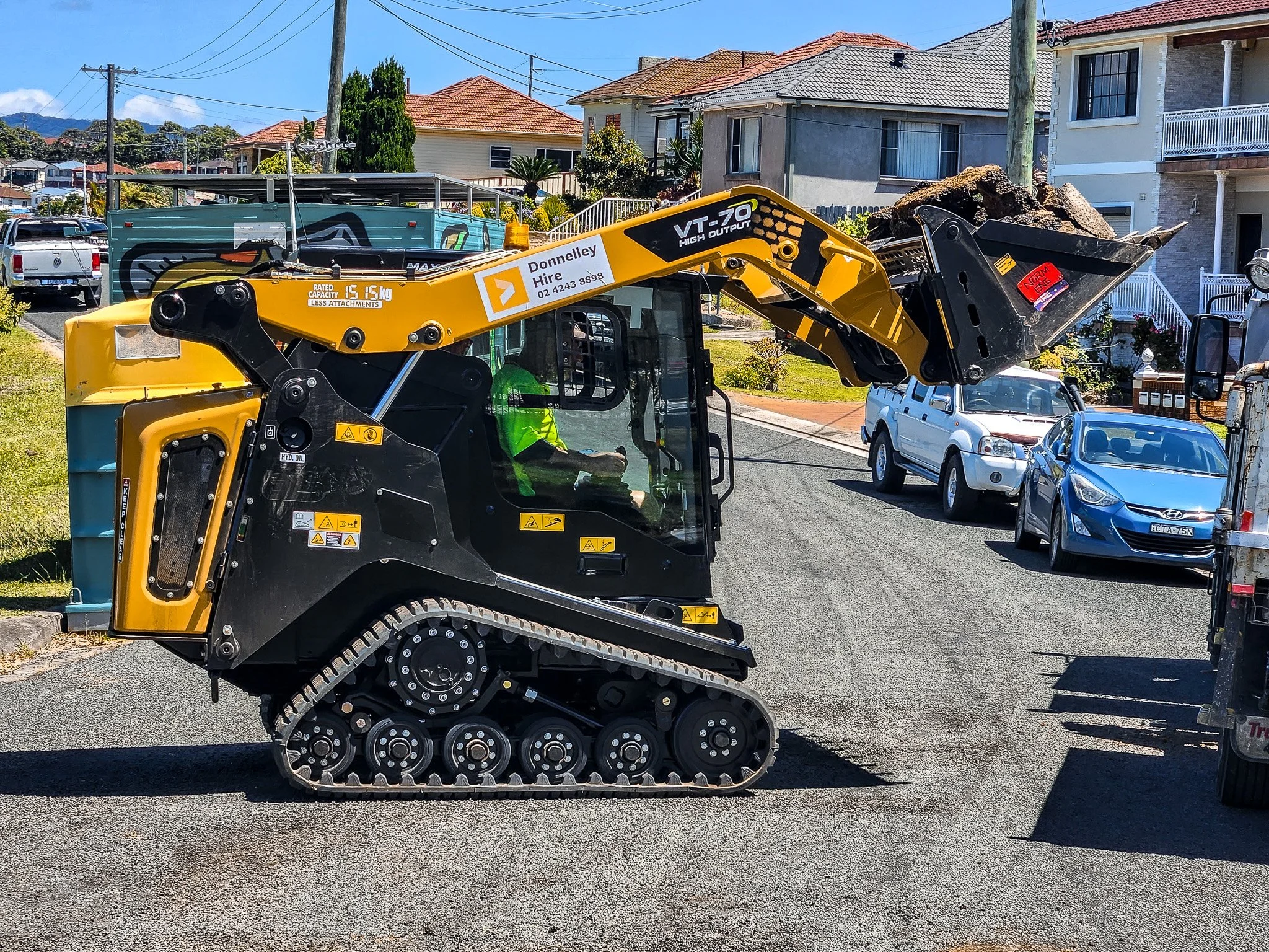 A small tracked skid steer loader lifting a load of dirt and rocks on a residential street with houses and parked cars in the background.