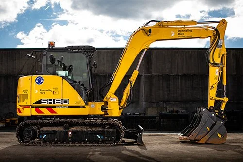 Yellow compact excavator with a digging bucket, parked outdoors against a black fence under cloudy sky.