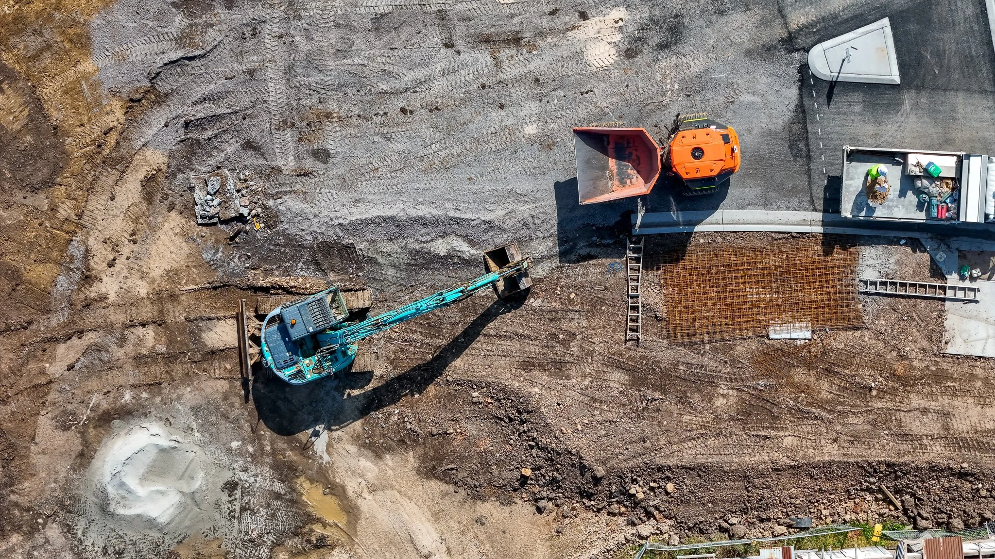 An aerial view of a construction site showing a blue excavator, orange construction vehicle, construction workers, and laying rebar on the ground.