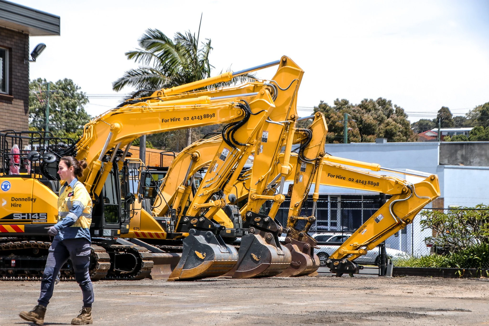 Several yellow excavators lined up at a construction site, with a woman walking past in the foreground.