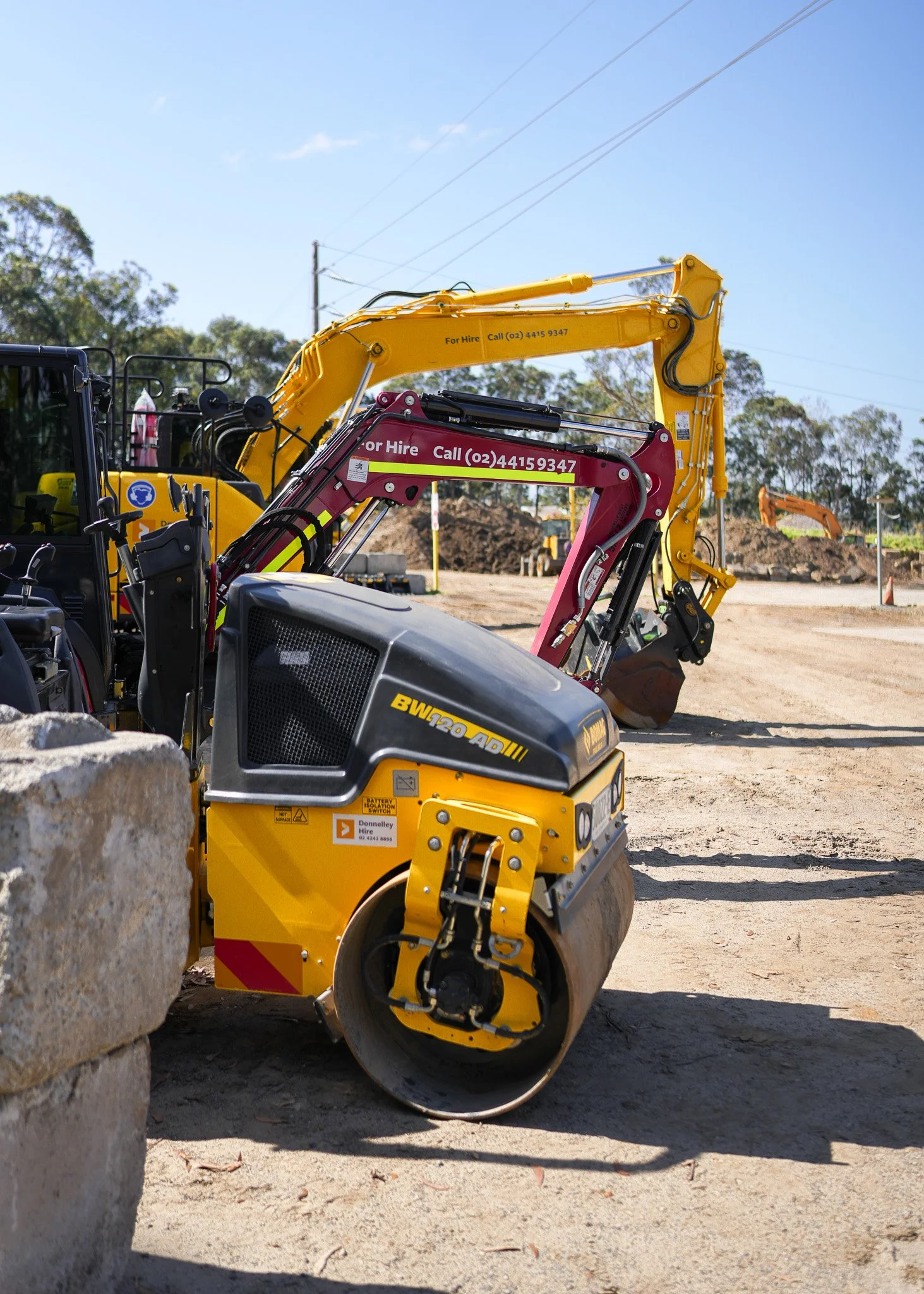 Construction site with yellow and pink excavators and a road roller in a clear sunny day.