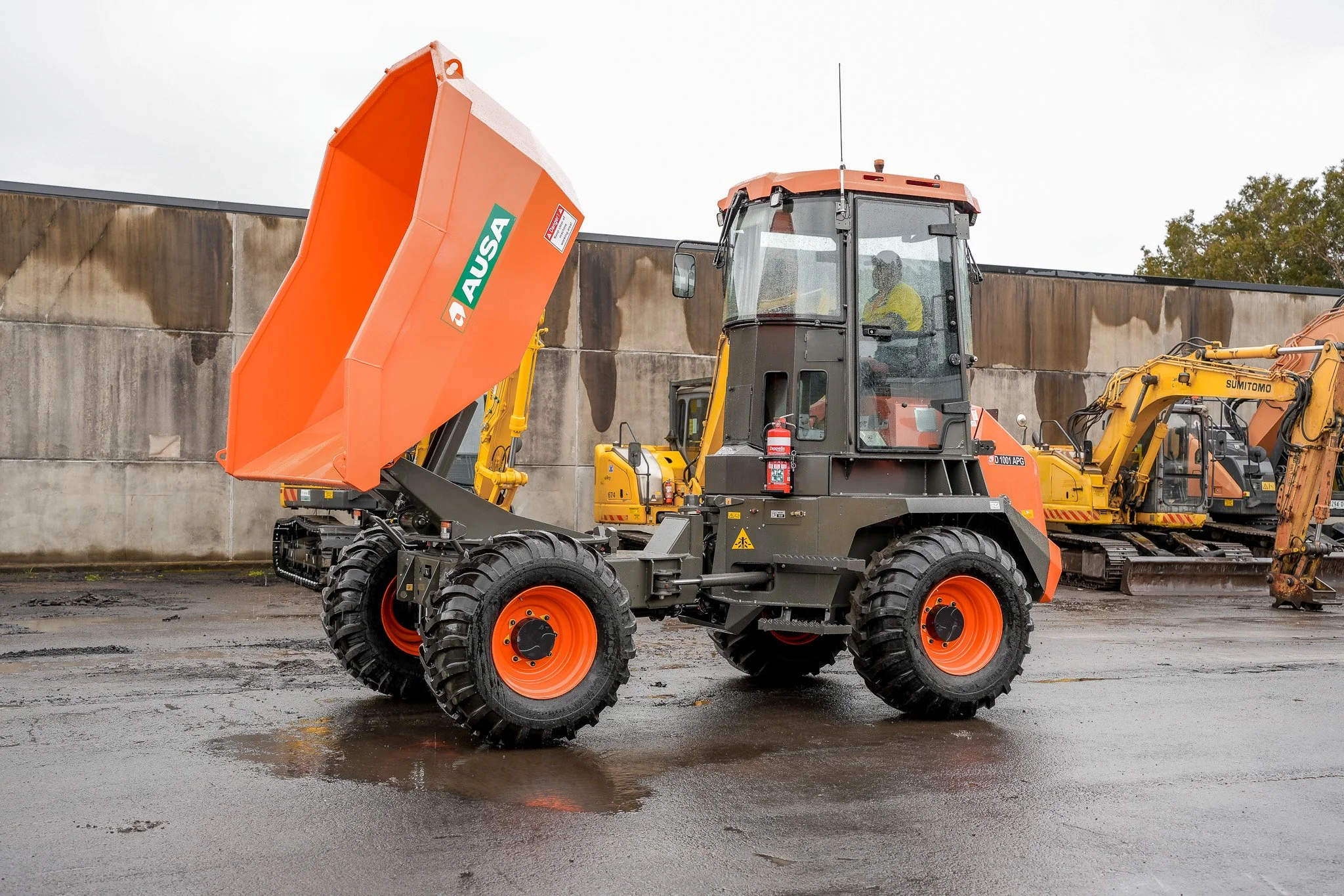 Construction vehicle with a raised orange dump bed, black and orange wheels, and a black cab, with other construction equipment in the background on a wet surface.