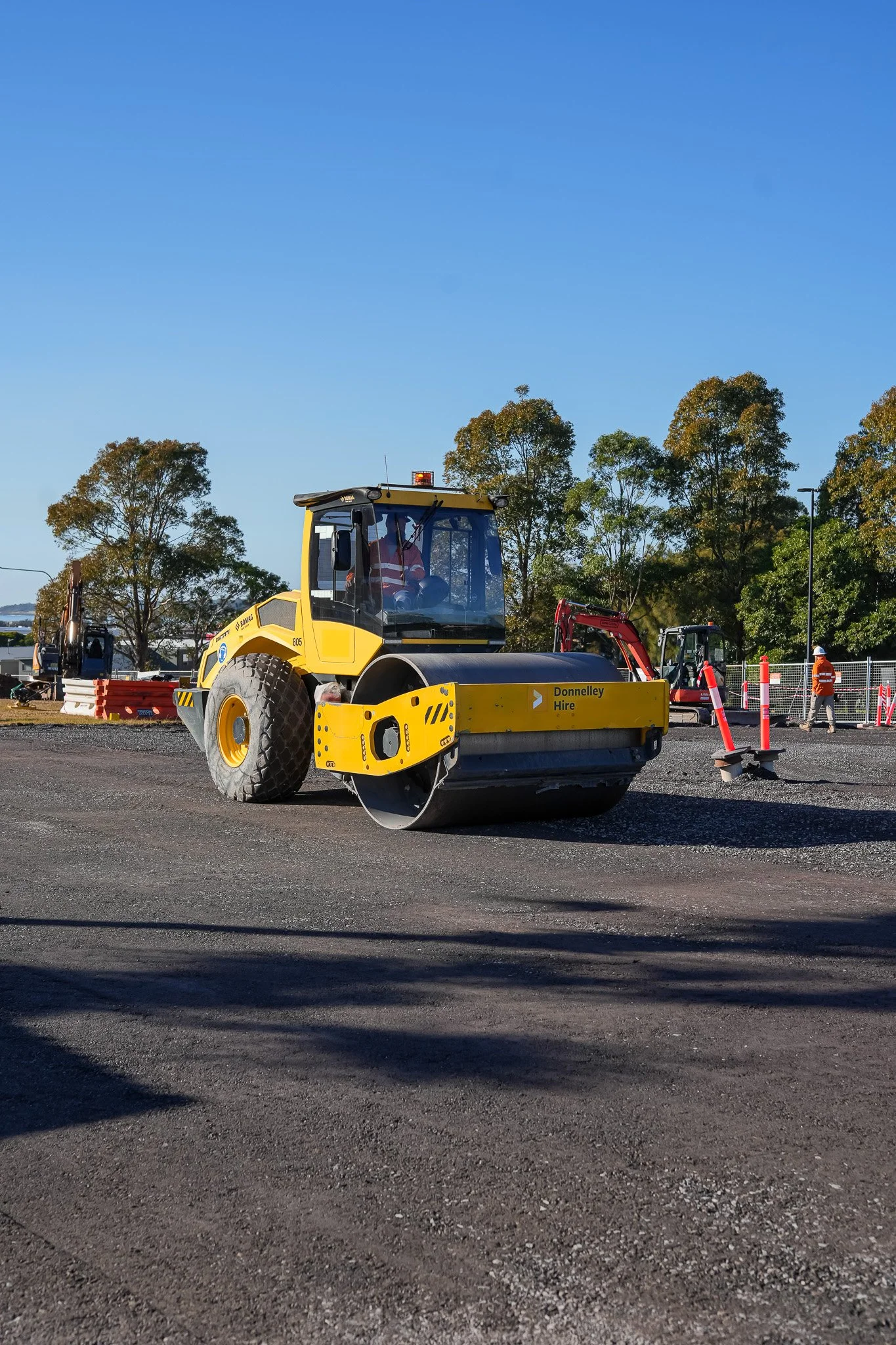A yellow road roller on a construction site with workers and trees in the background.