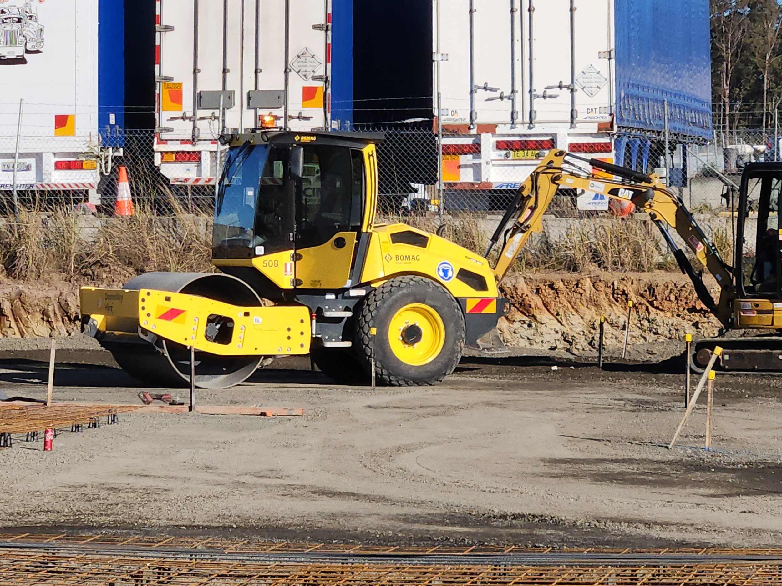 Construction site with a yellow soil compactor and a small excavator, with trucks in the background.