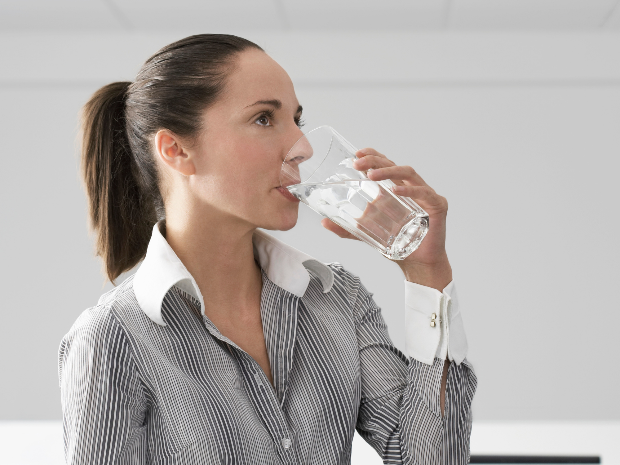 Woman with brown hair tied in a ponytail wearing a striped shirt drinking a glass of water in an indoor setting.