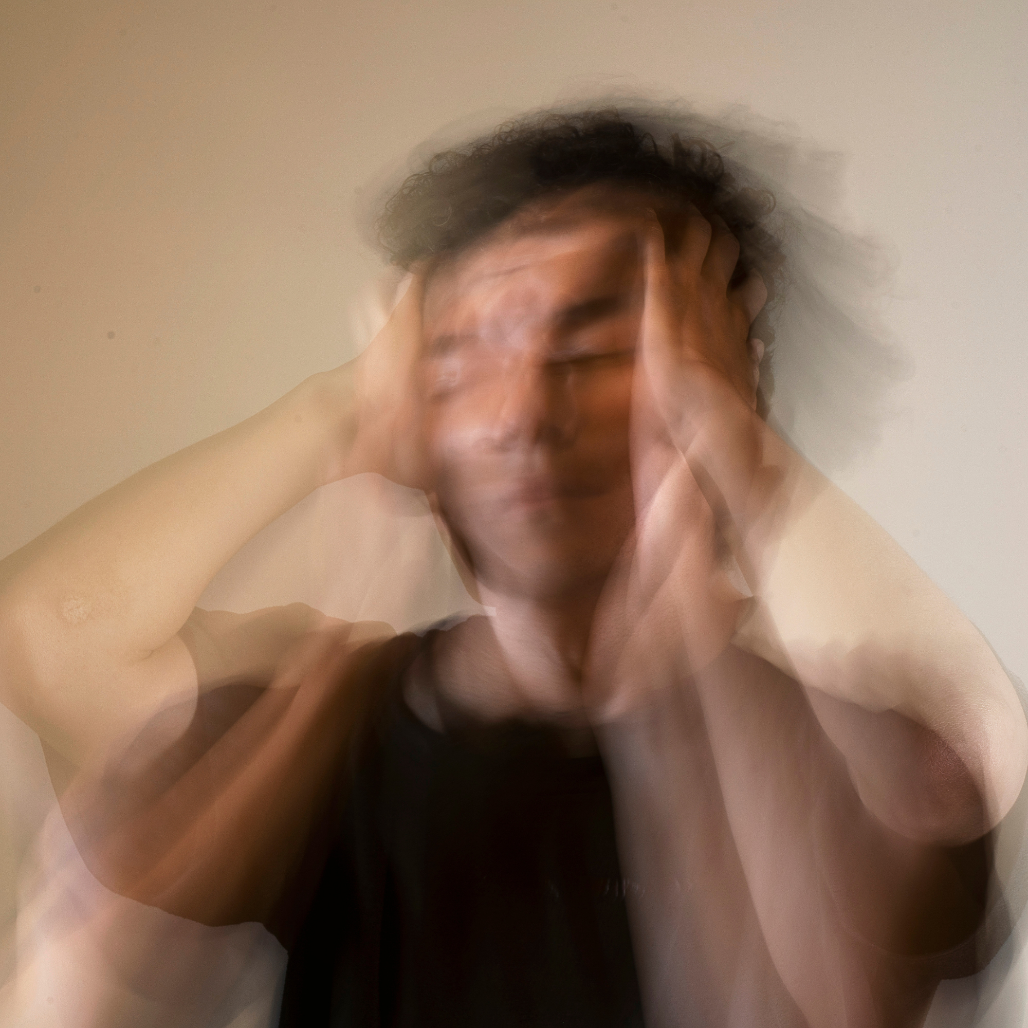 A person with curly hair holding their head with both hands, displaying a distressed expression, with a blurred, multi-exposure effect.