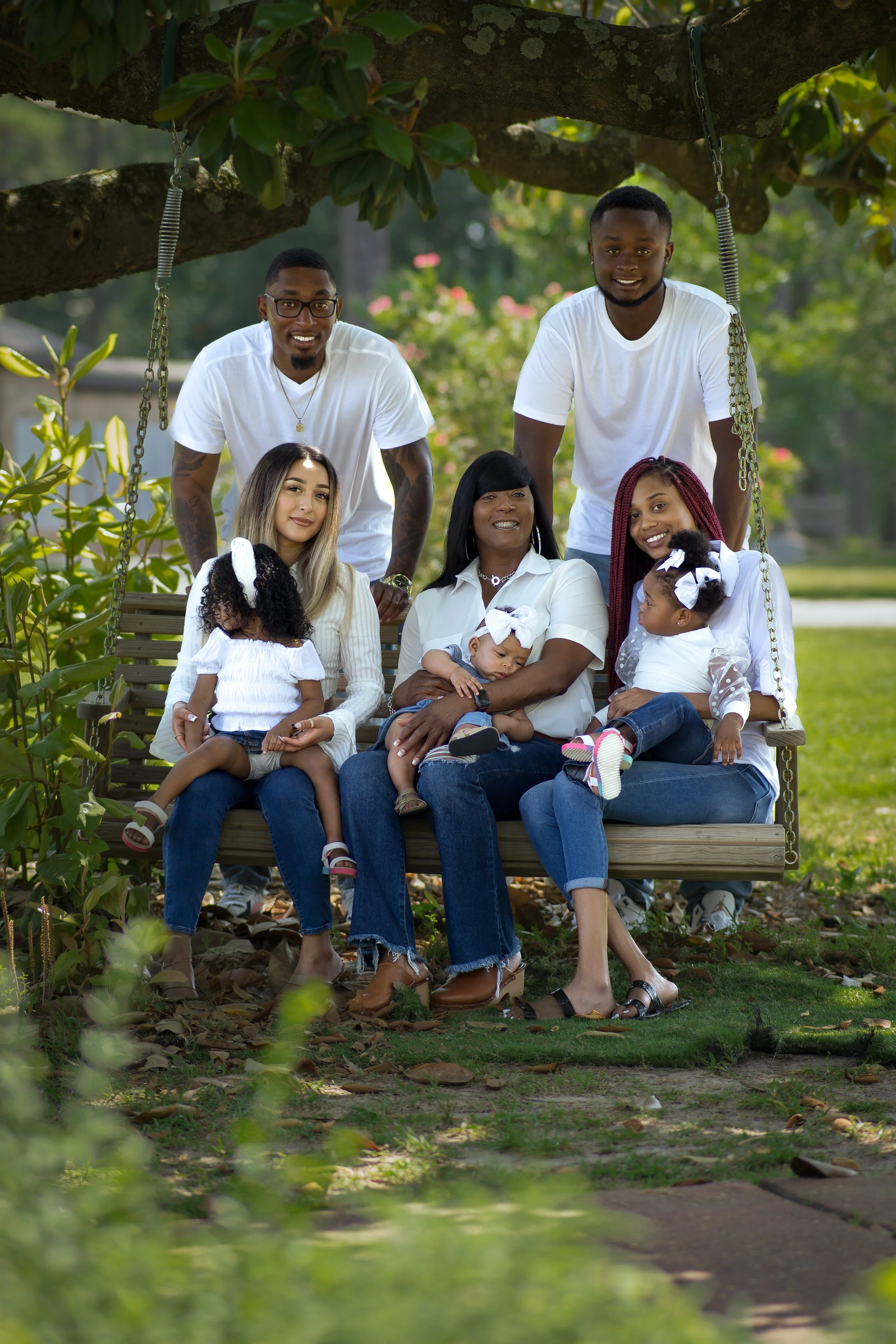 Family photo of seven people, including three adults and four children, sitting on a wooden swing under a tree in a park.