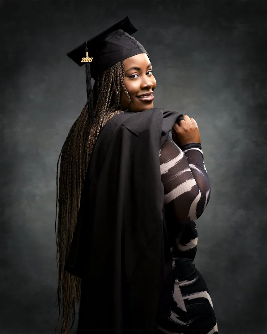 A woman in a black graduation cap and gown looking over her shoulder, smiling, with long braids, against a dark background.