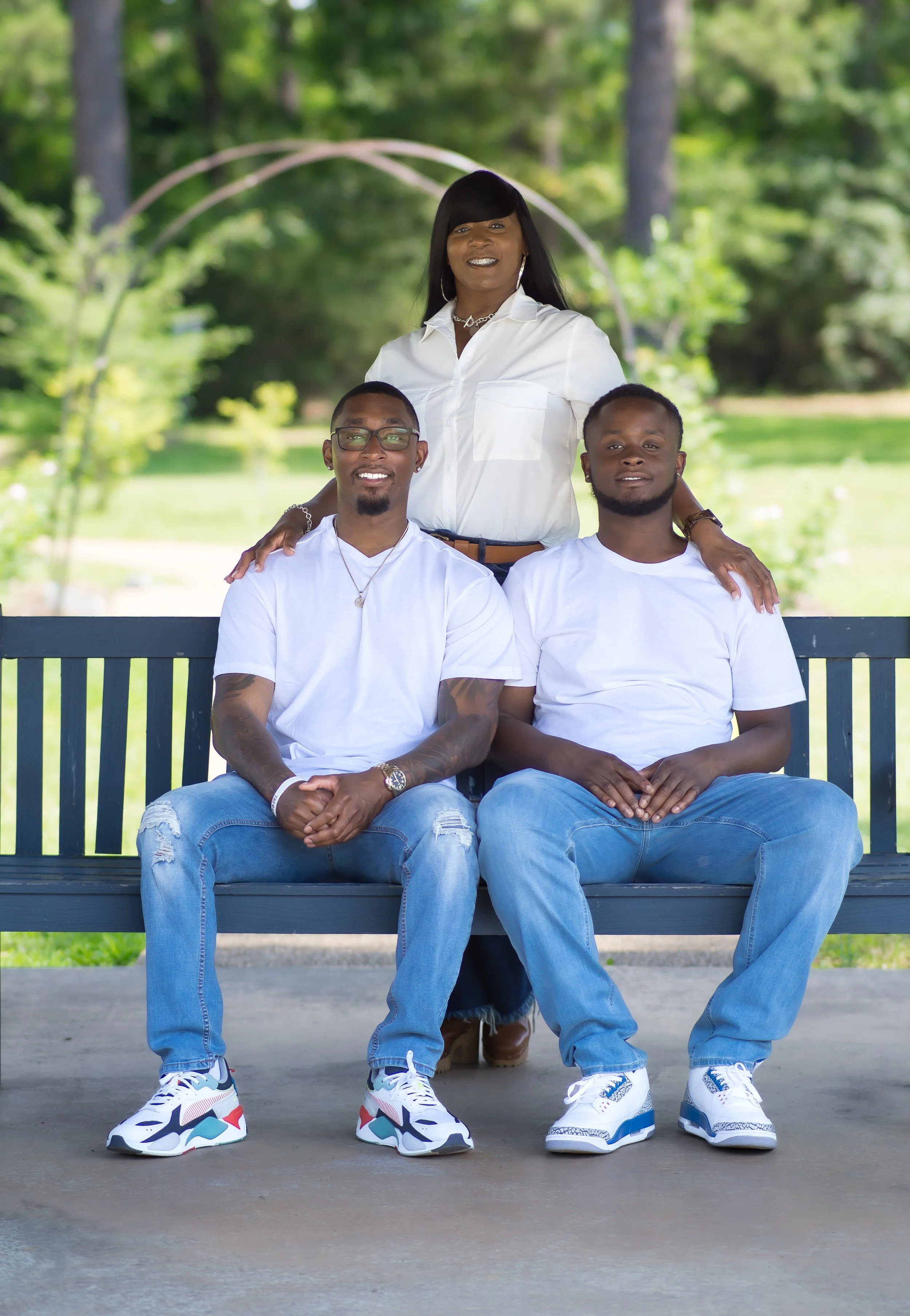 A family of three posing in a park on a bench, with a woman standing behind two men, all smiling, dressed casually in white shirts and jeans, with greenery in the background.