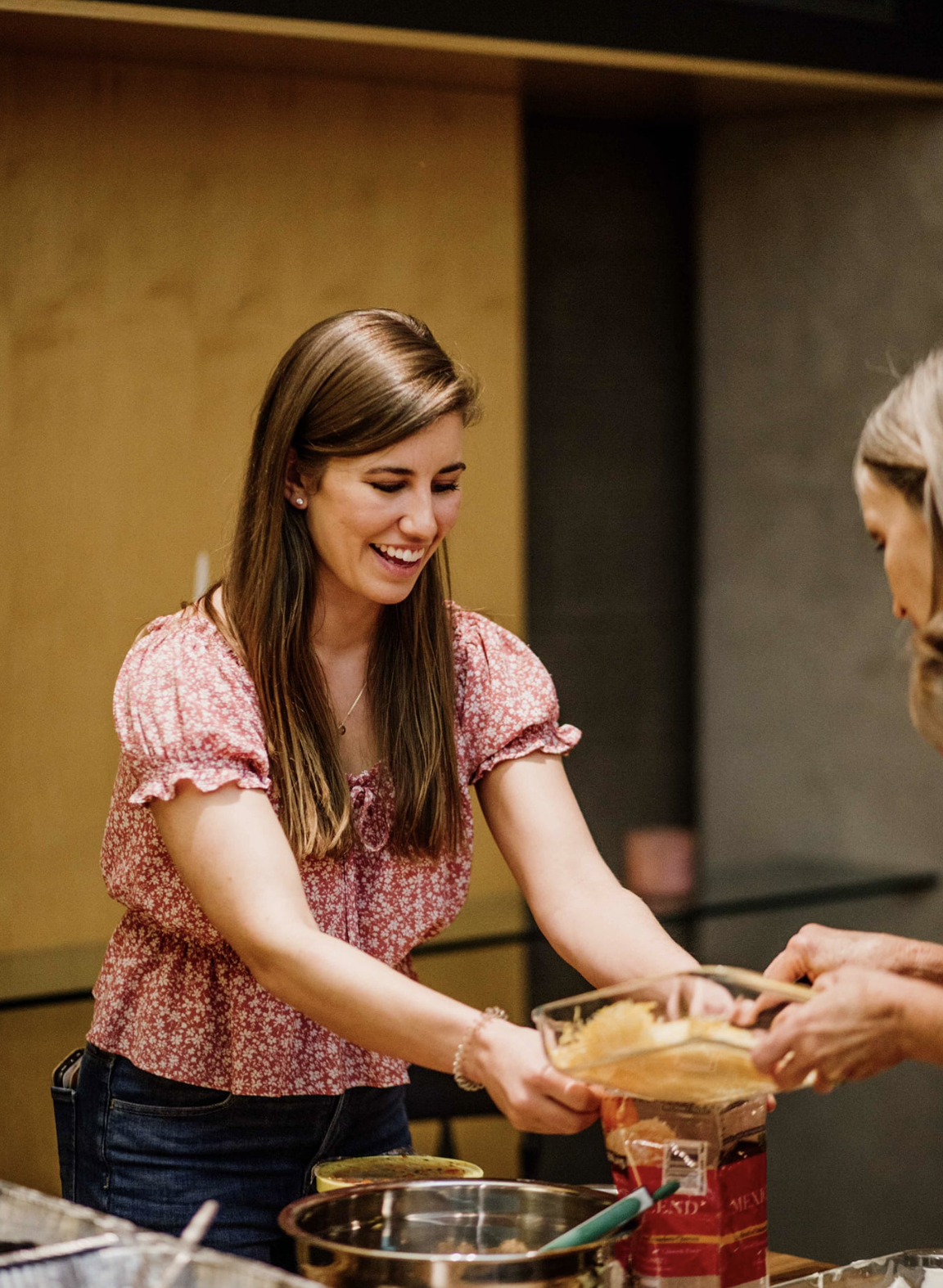 A woman in a pink floral blouse is smiling and serving food at a table, engaging in a friendly interaction.