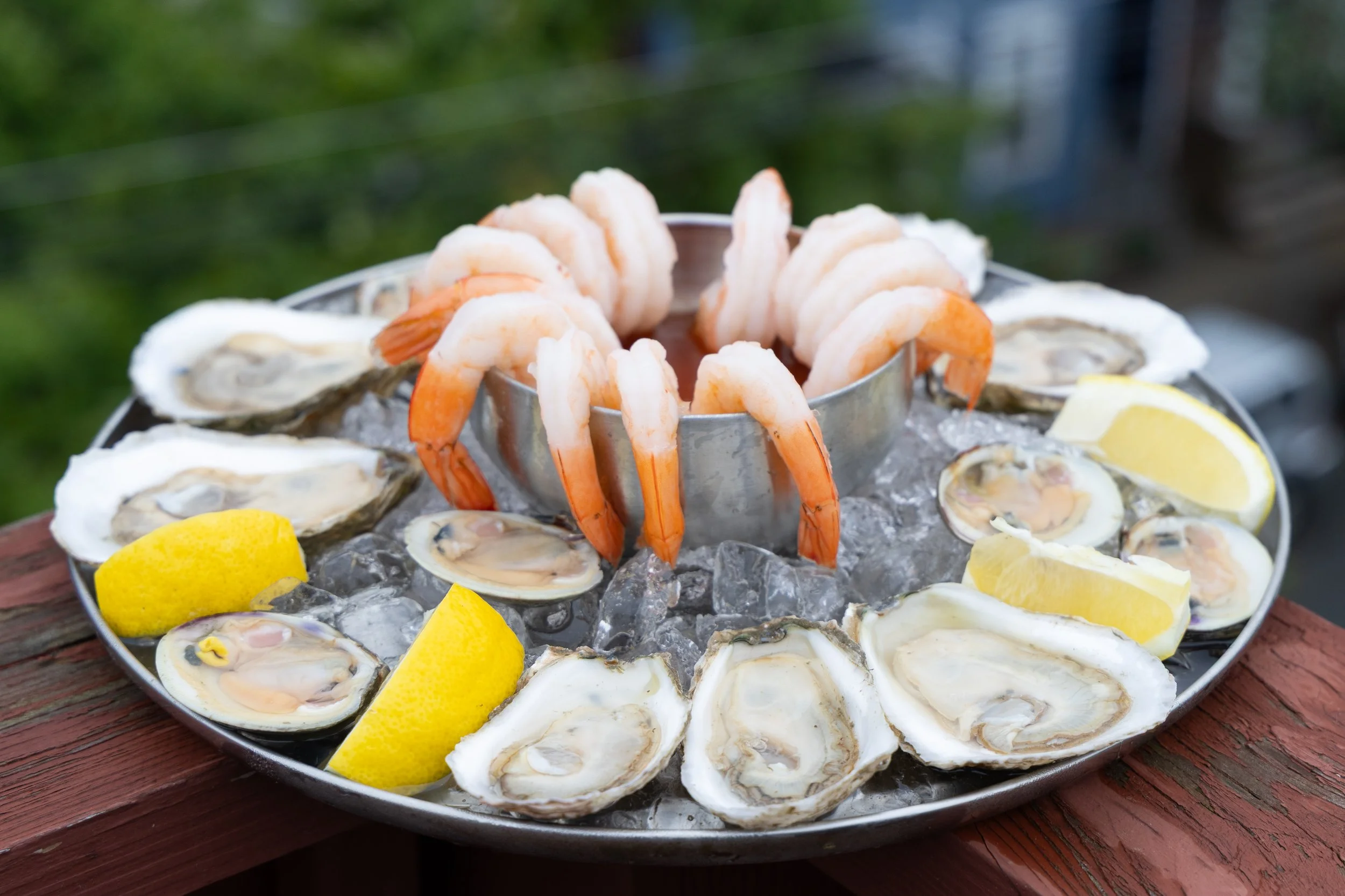 A round tray of fresh oysters on the half shell with lemon wedges on ice, topped with shrimp, outdoors on a wooden surface. oyster bar boston new england